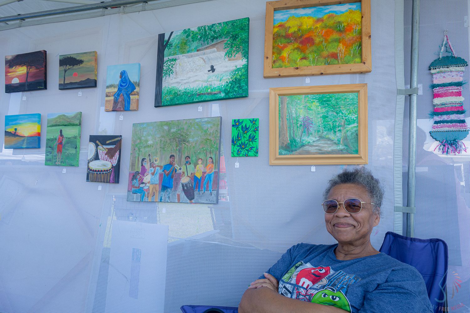 Elderly woman sitting in an art booth surrounded by colorful paintings displayed on a white wall at an outdoor art fair.