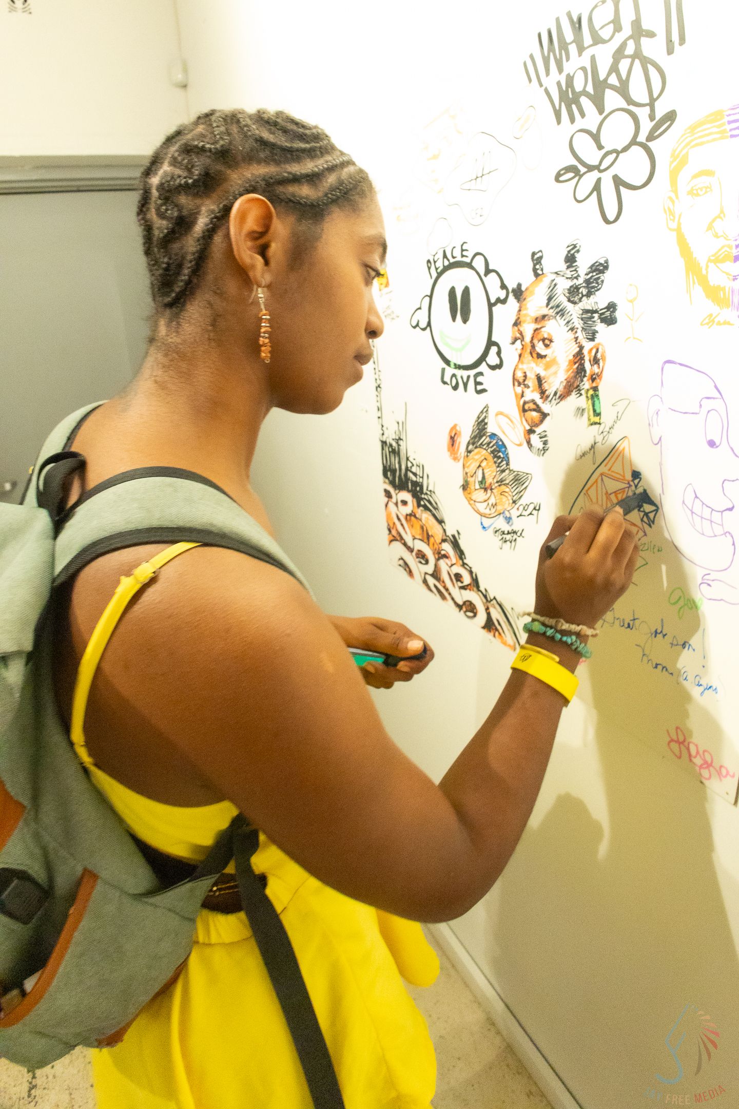 Young woman with braids and yellow dress draws on a white wall covered in colorful doodles and messages.