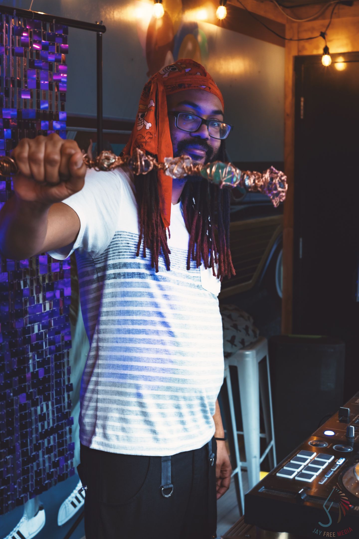 Man with red dreadlocks and glasses holds a skewer in a dimly lit bar with DJ equipment nearby.