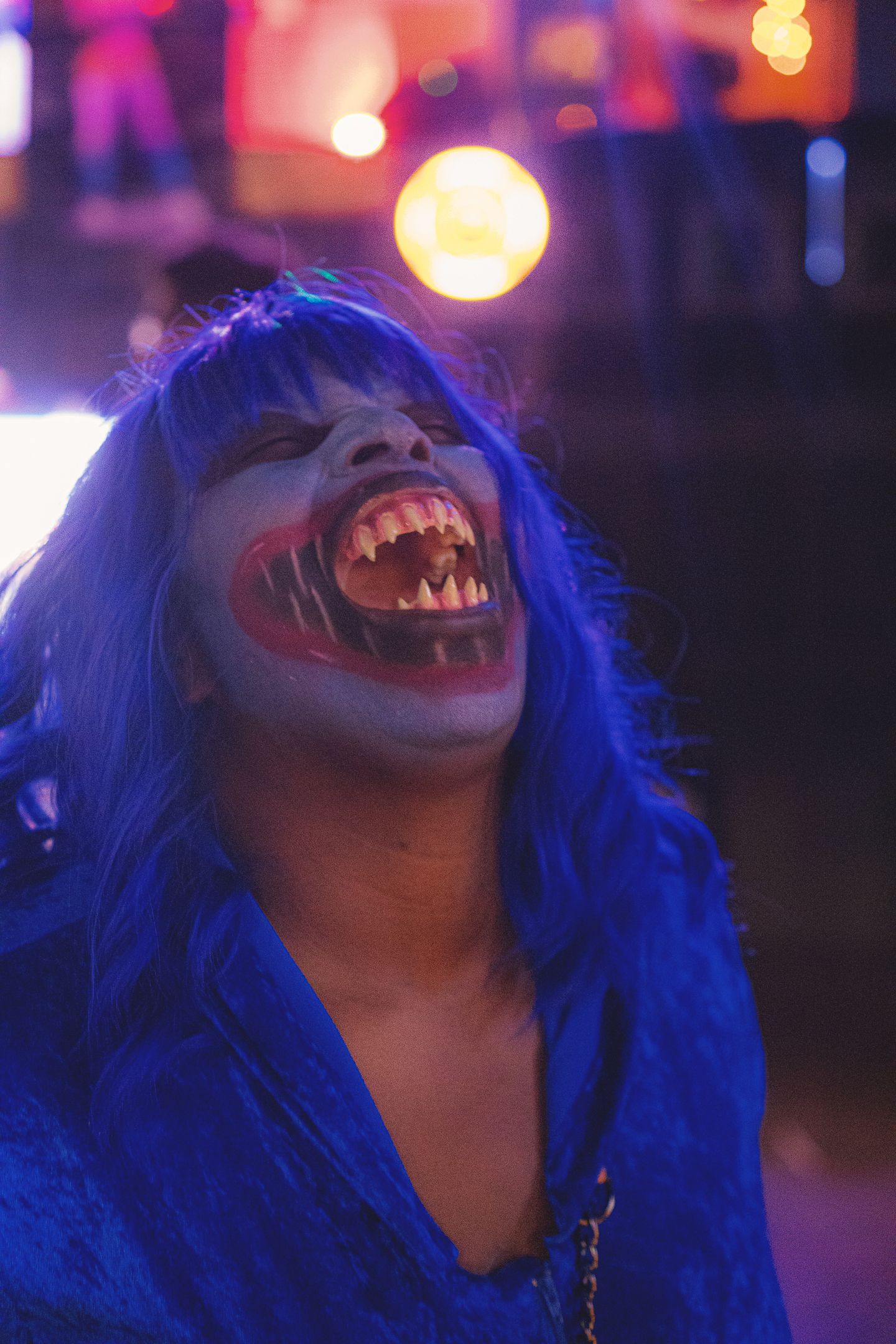 Person in vampire Halloween costume with blue hair, fangs, and dramatic makeup under purple neon lights.