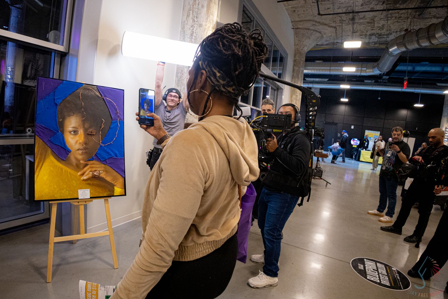 Art gallery event with a colorful portrait painting on an easel, attendees and a camera crew in the background.