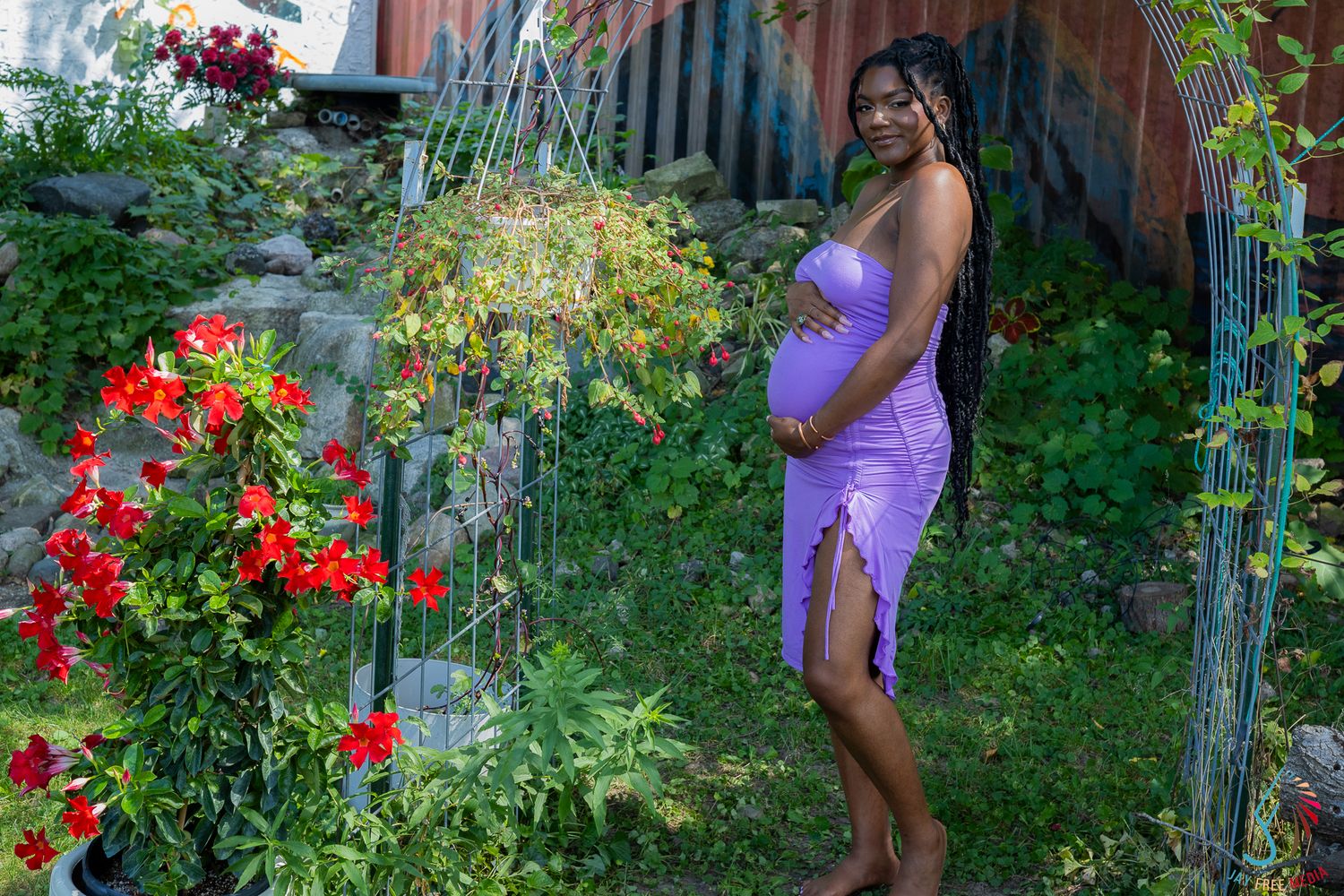 Pregnant Black woman in a purple dress posing in a lush garden surrounded by red flowers and greenery.