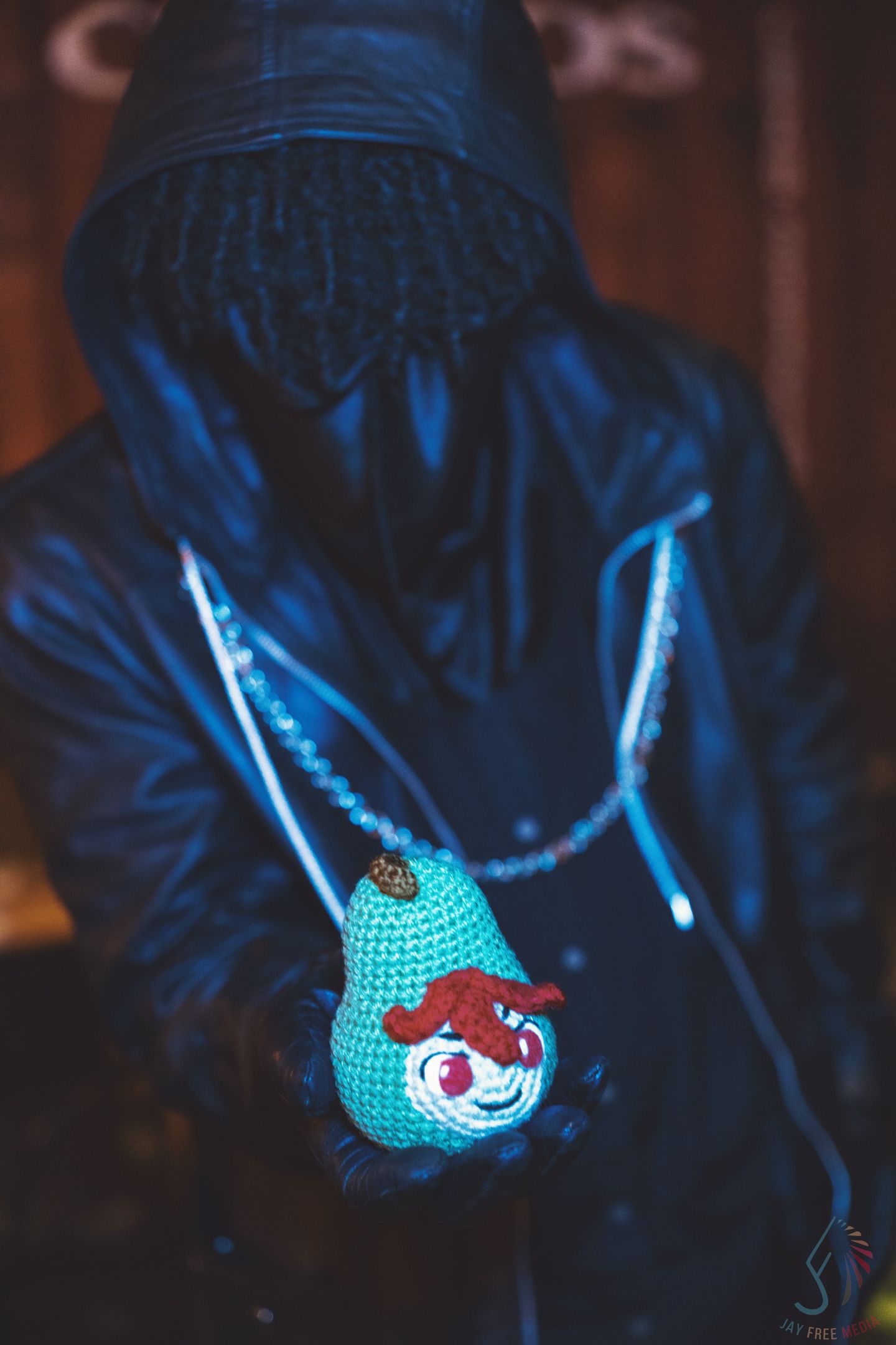Mysterious cosplayer in black hooded jacket holds a teal crocheted clown doll accessory at night.