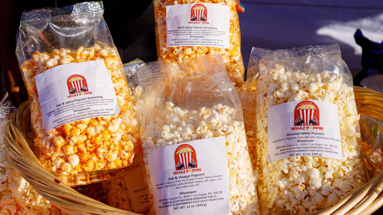 Bags of fresh popcorn with branded labels displayed in a wicker basket at an outdoor market or festival.