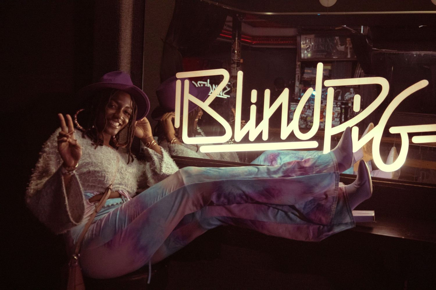 Woman posing playfully in front of a Blind Pig neon sign, wearing a purple hat and colorful tie-dye pants.