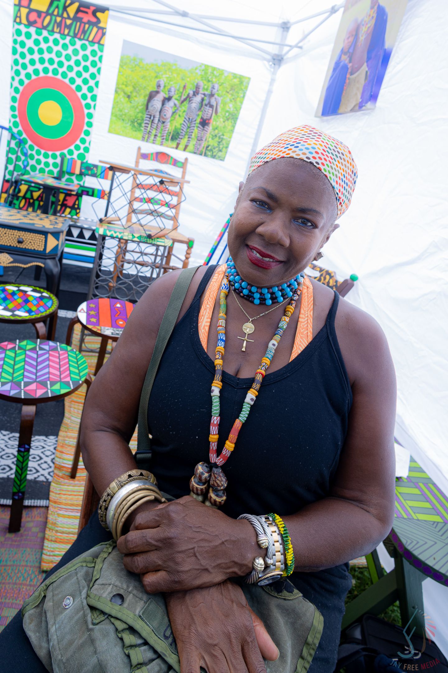 A woman wearing colorful beaded jewelry and a patterned headscarf smiles at an outdoor art fair.