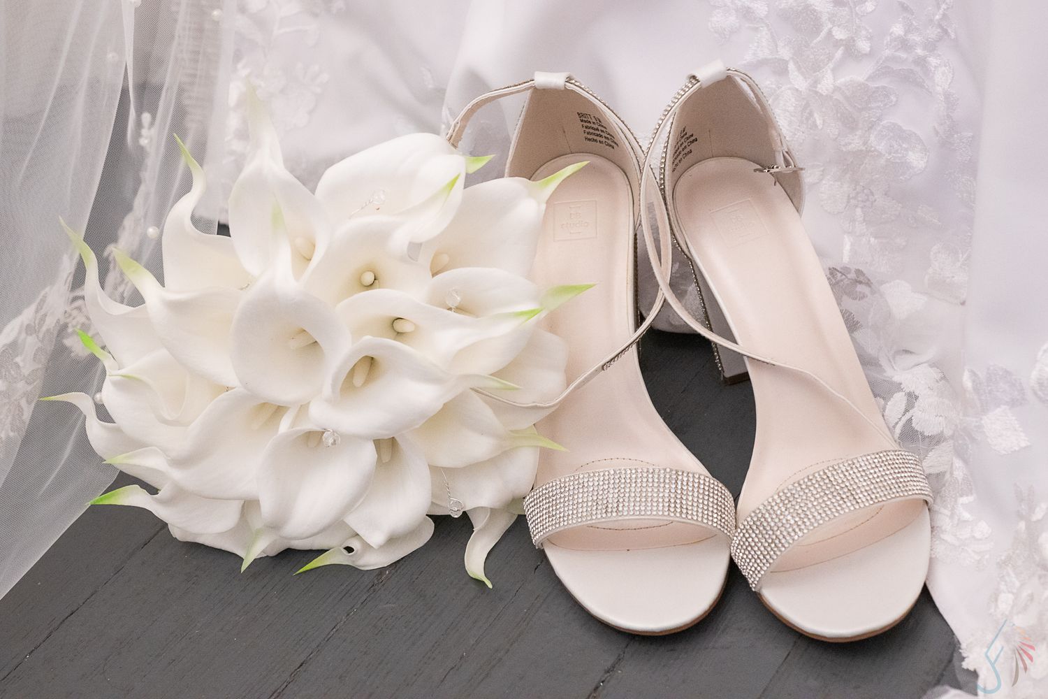 Elegant bridal flat sandals with rhinestone embellishment beside white calla lily bouquet on gray surface.
