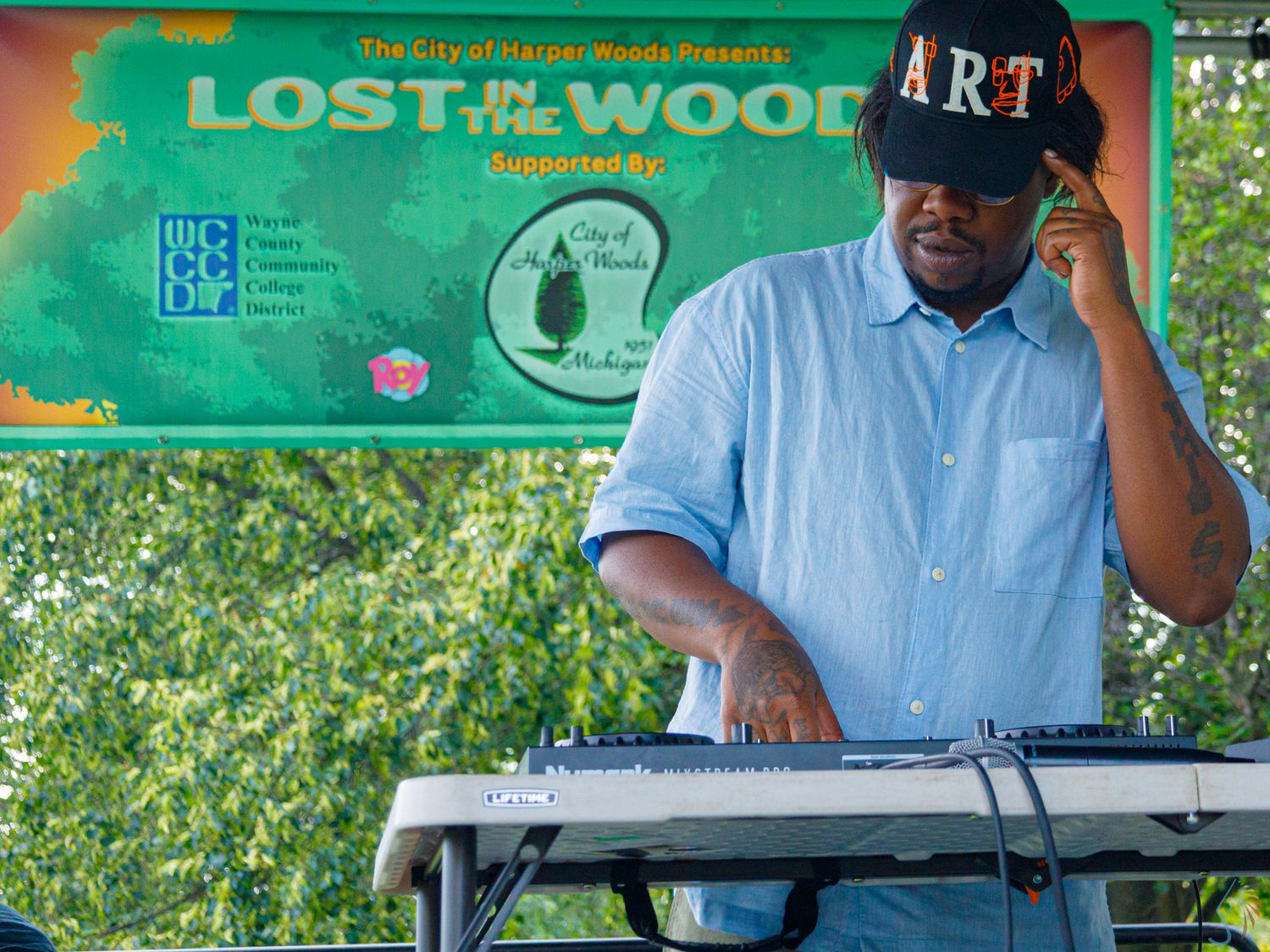DJ performing on keyboard at Lost in the Woods outdoor music festival, wearing ART cap, green banner background.
