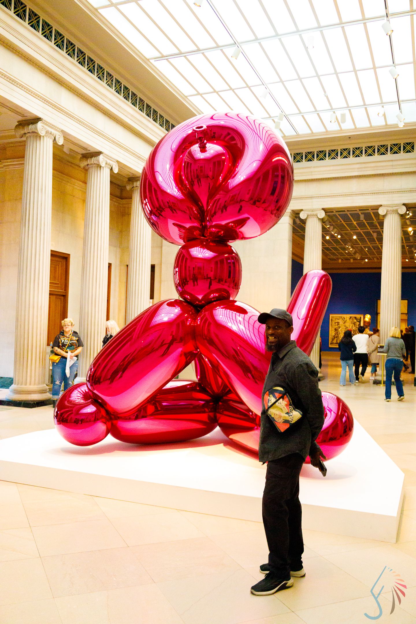 Man posing in front of a large shiny red balloon dog sculpture inside a grand museum hall with columns.