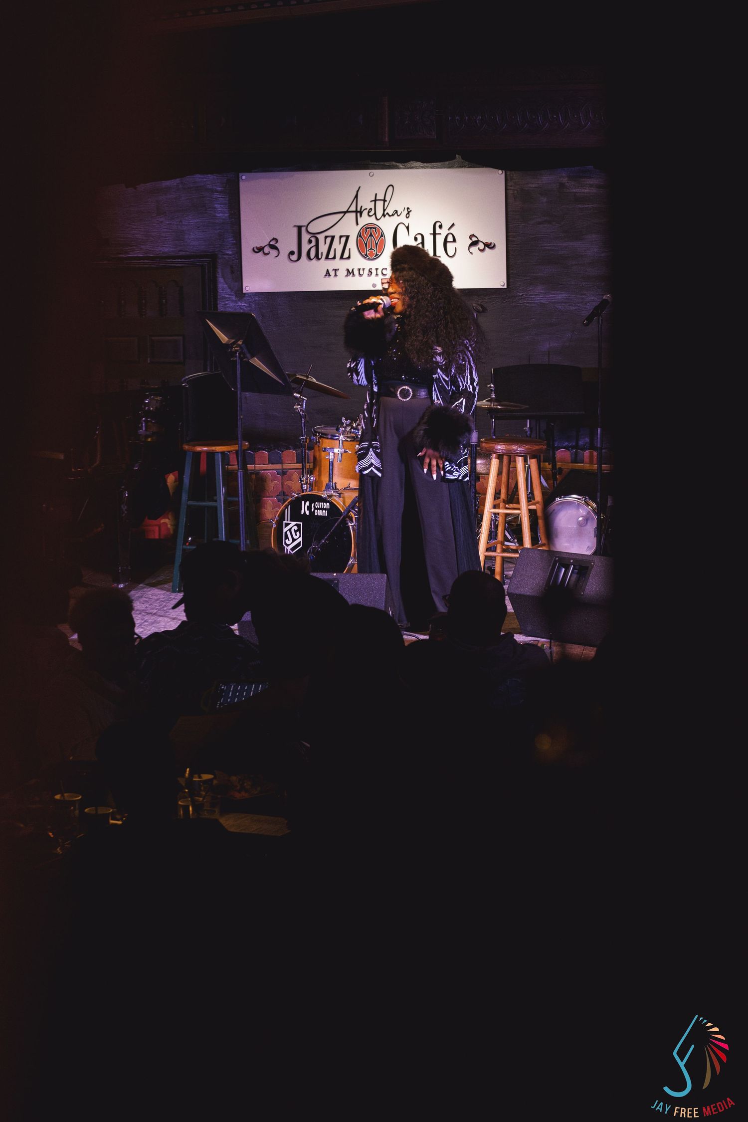 Live jazz performance at Jazz Cafe, singer on dimly lit stage with drum kit and bar stools in background.