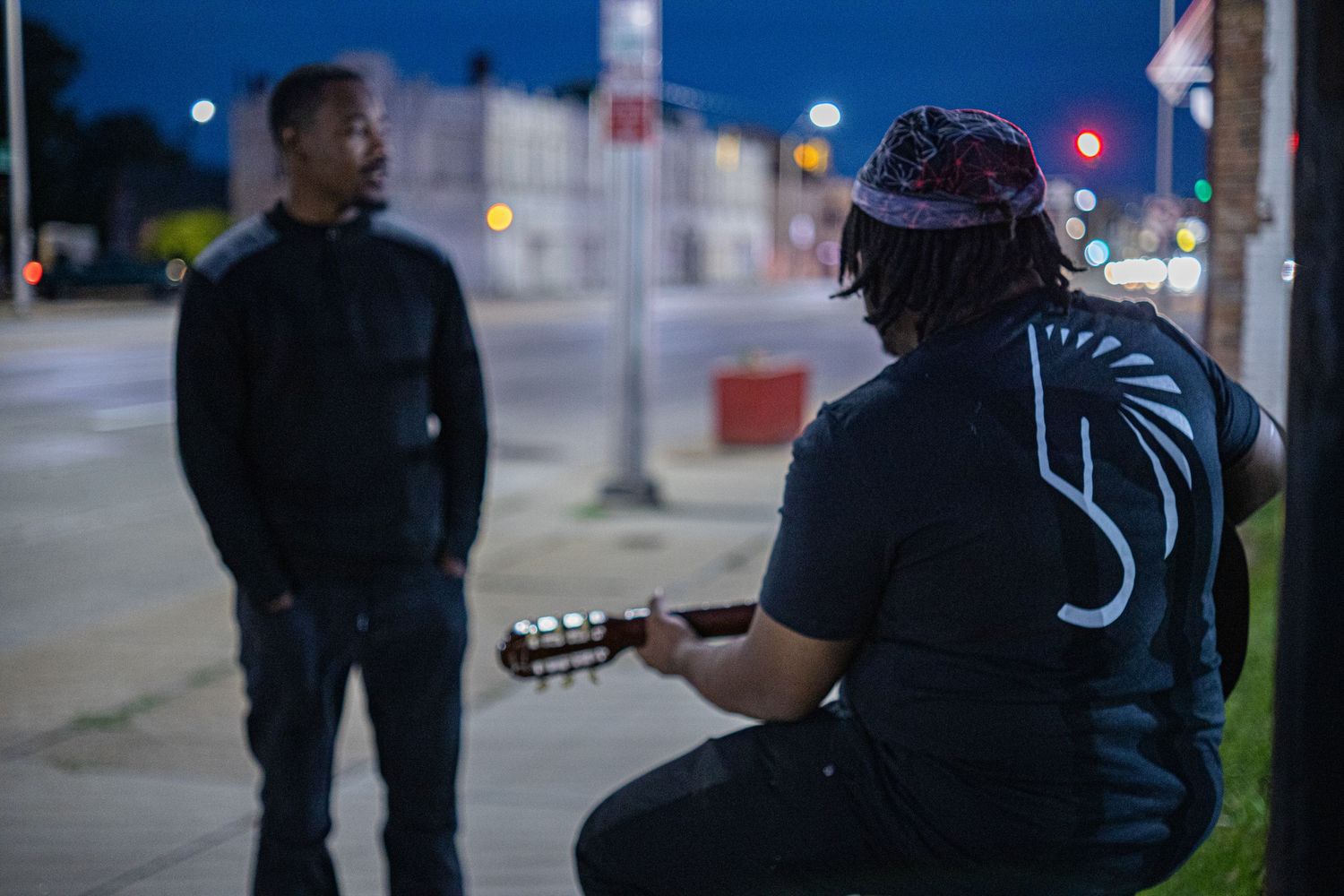Musician plays guitar outdoors at night while another person watches on city street.