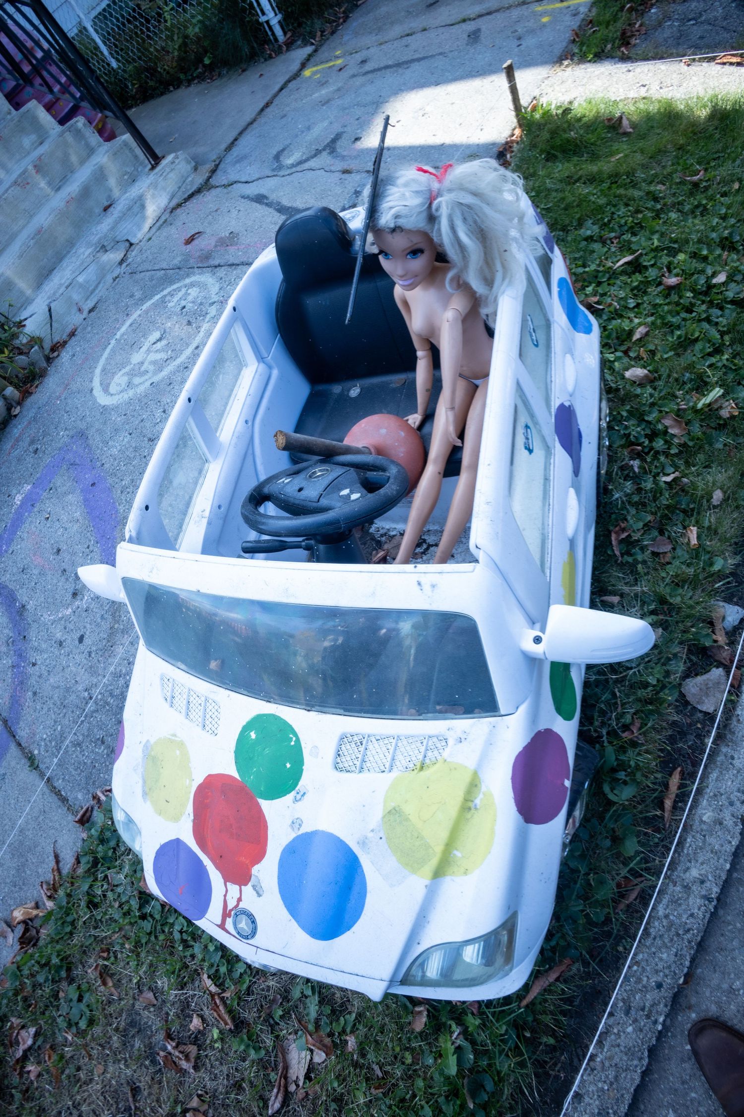 A naked Barbie doll sits in a worn white ride-on toy car decorated with colorful balloon designs on a sidewalk.