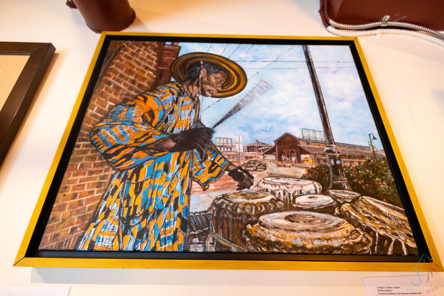 Colorful painting of an African musician in traditional attire playing near drums, displayed in a gold frame.