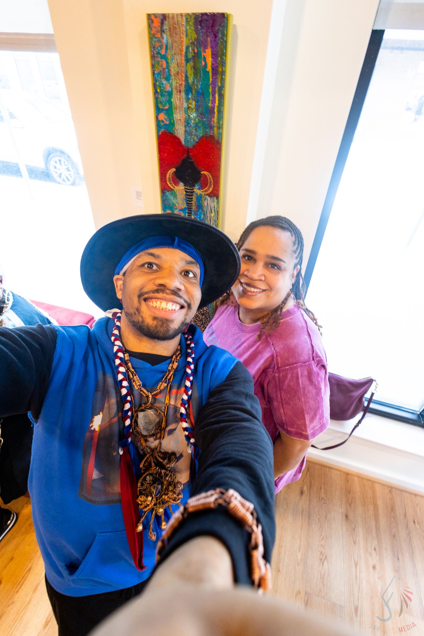 A smiling man in a blue shirt and hat takes a selfie with a woman in pink inside an art gallery.