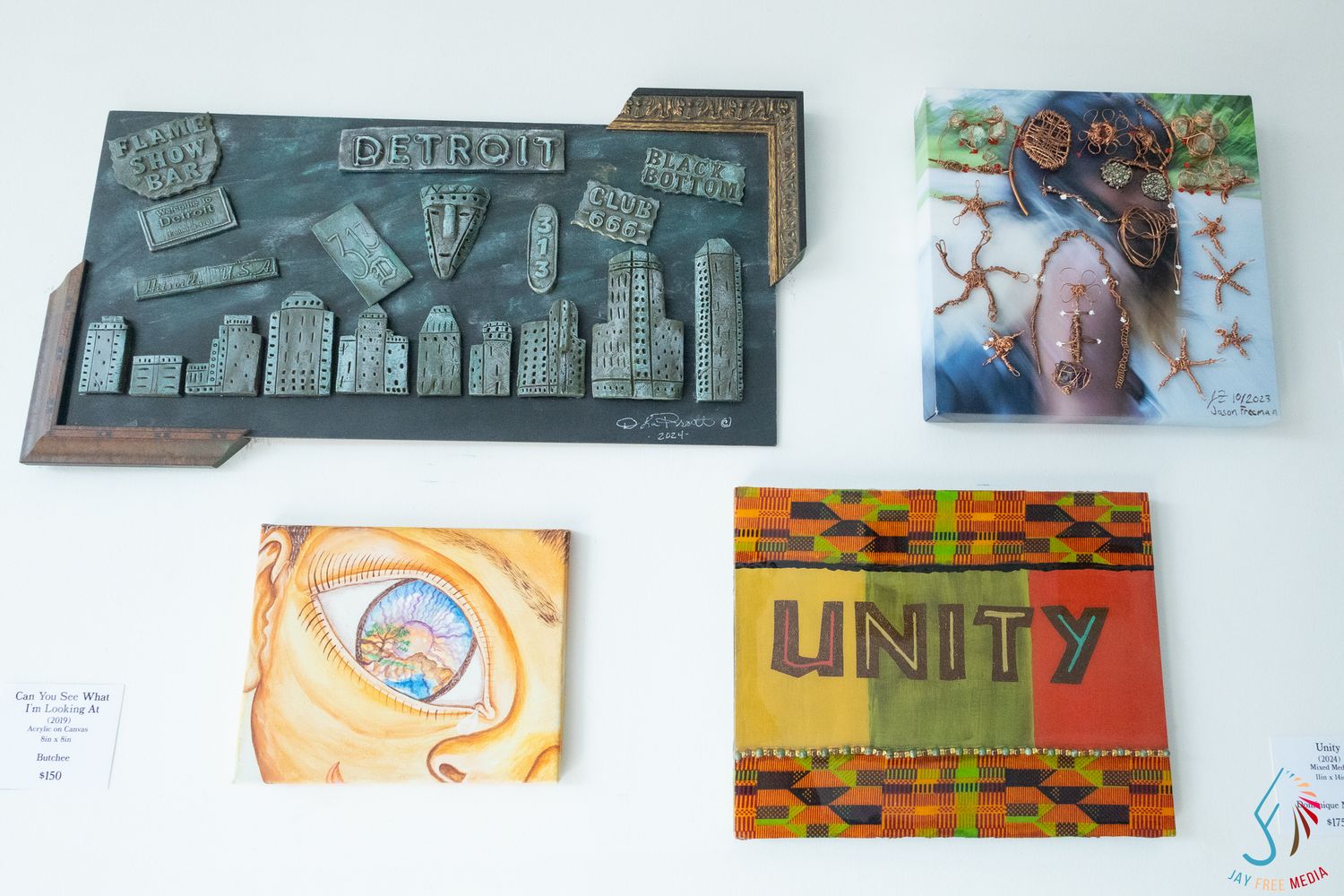 Four pieces of artwork displayed on a white wall, including a Detroit cityscape relief, Unity sign, and two colorful paintings.