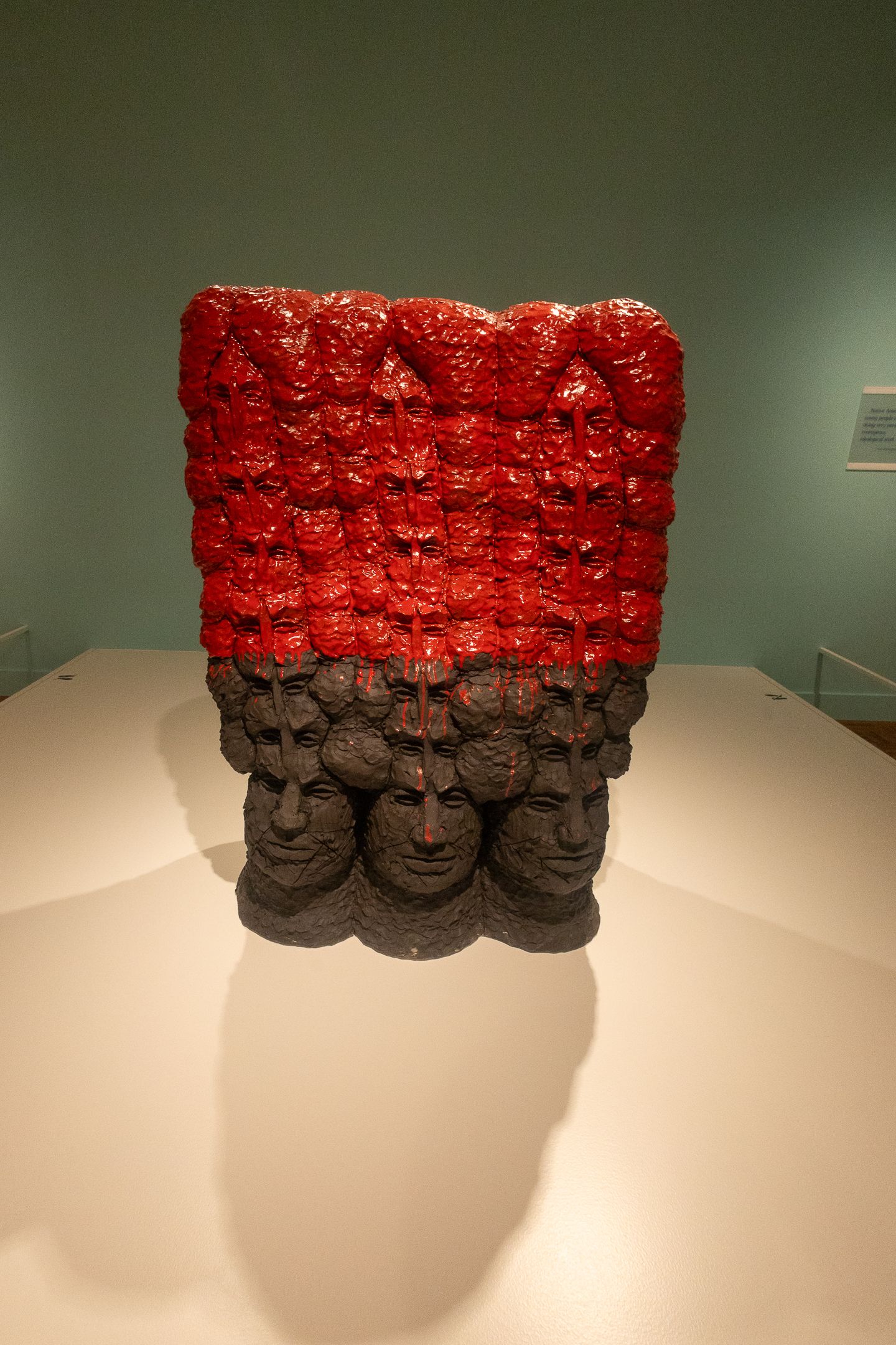 Sculpture of stacked human faces in dark stone base topped with vibrant red figures on display in a museum.
