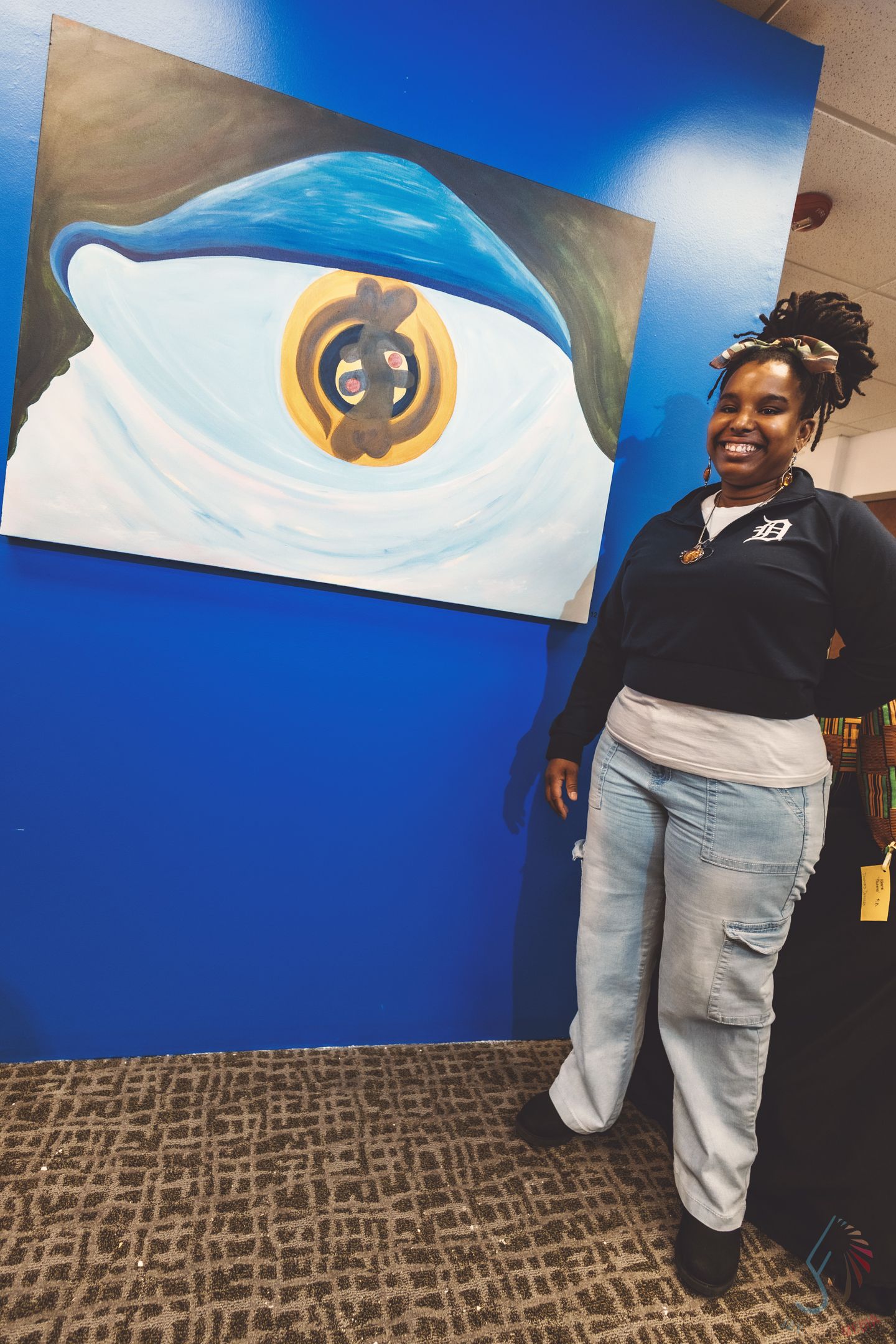 Woman smiling beside a large eye painting on a blue wall at an art gallery, wearing a Detroit shirt.