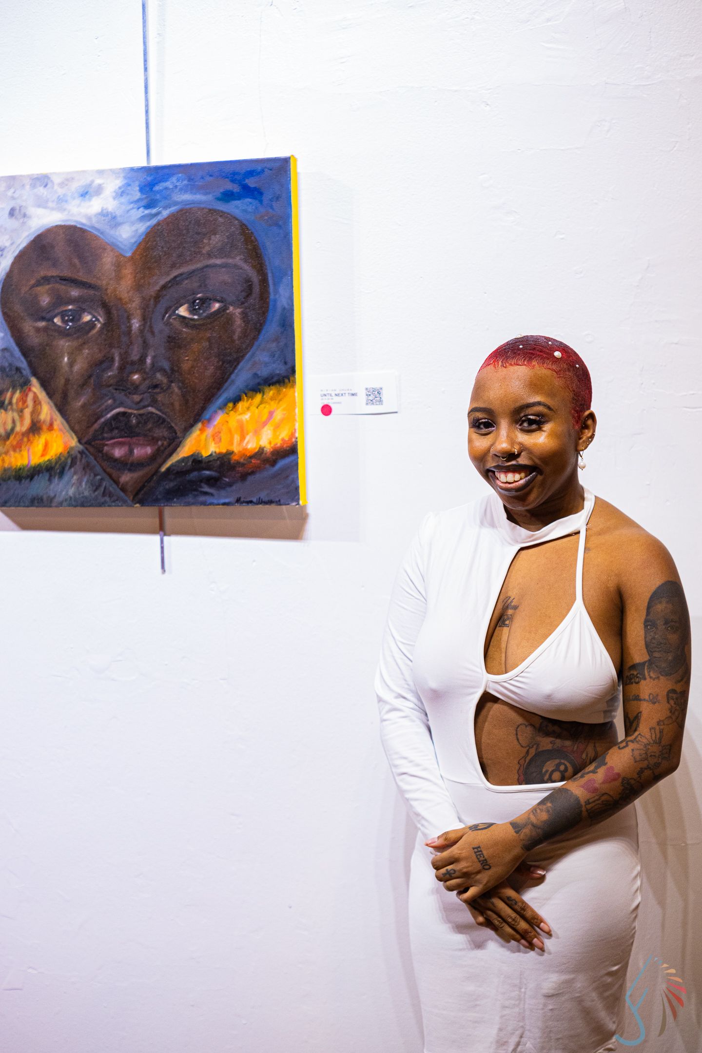 Smiling woman in white outfit with tattoos posing beside heart-shaped face painting at art gallery.