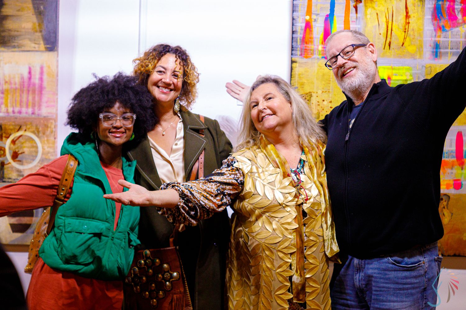 Four diverse adults smiling and posing together in a colorful art studio, one wearing a gold sequin jacket.