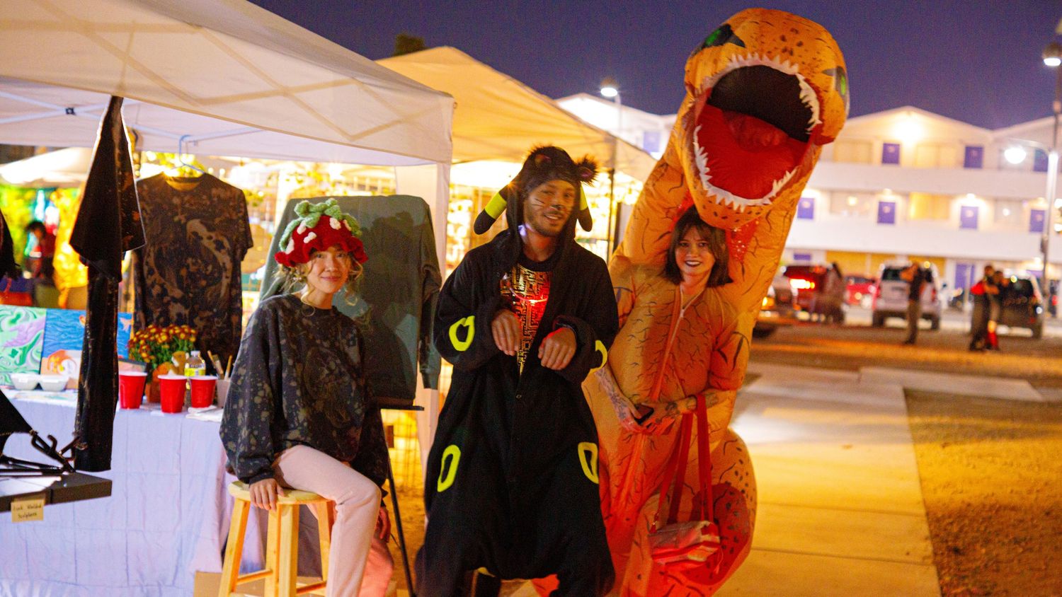 Three people in Halloween costumes including dinosaur and pirate at outdoor night market.