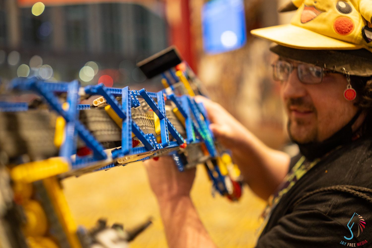 Man in Pikachu hat examining a blue LEGO creation at a convention display.