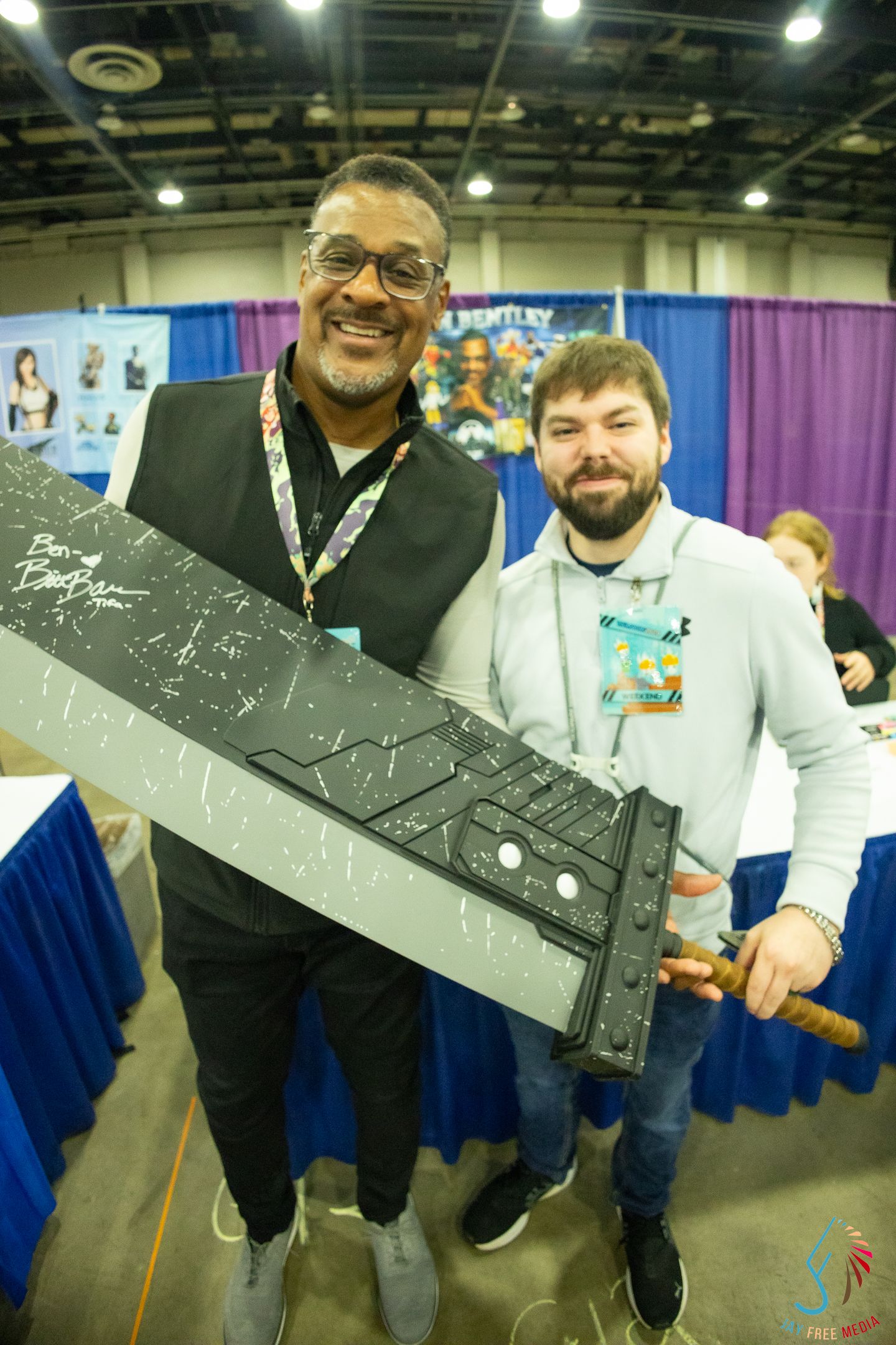 Two men smile holding large prop sword with signatures at comic convention vendor booth.