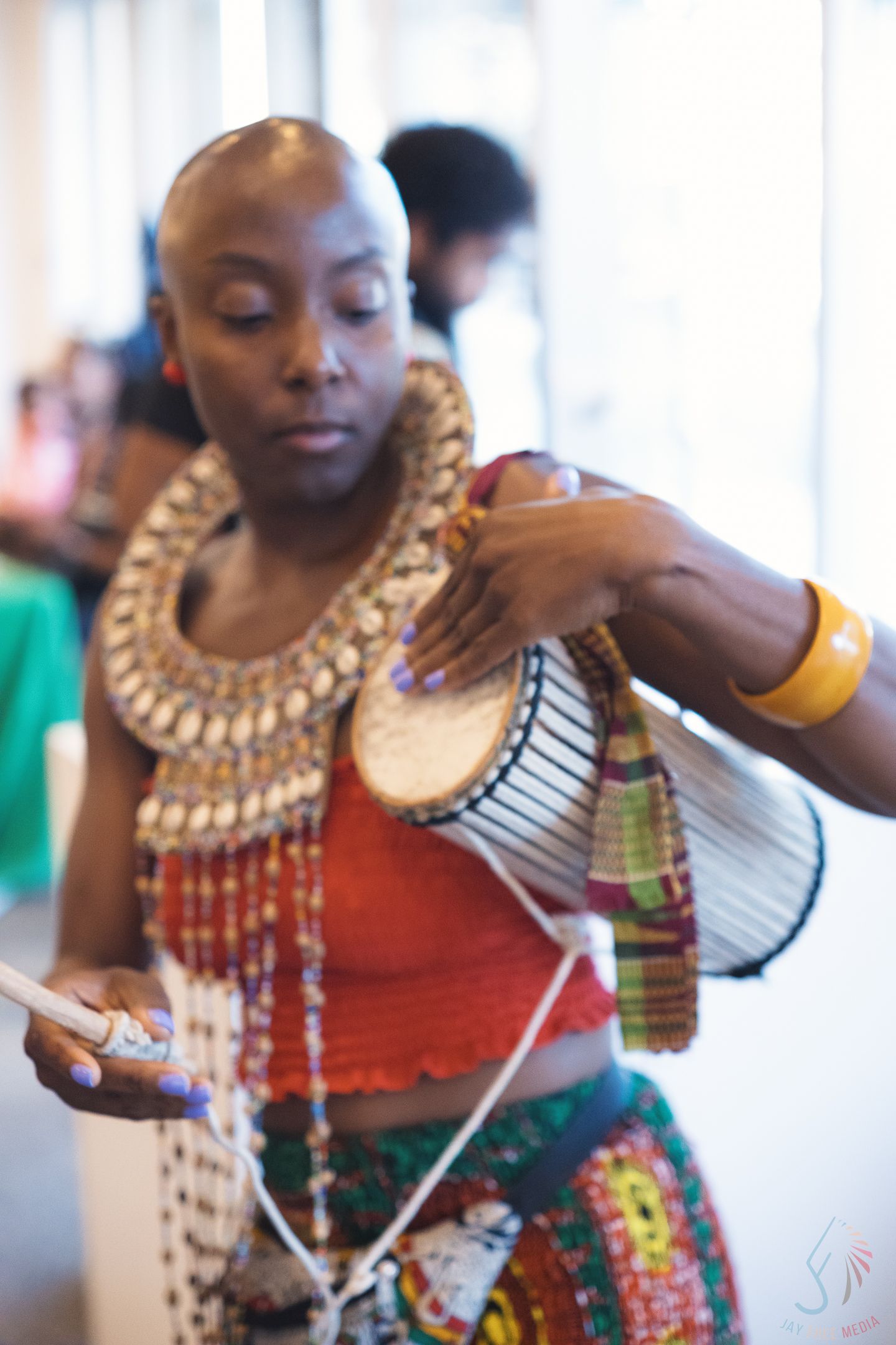 African woman in traditional beaded necklace and colorful attire playing a djembe drum at a cultural event.