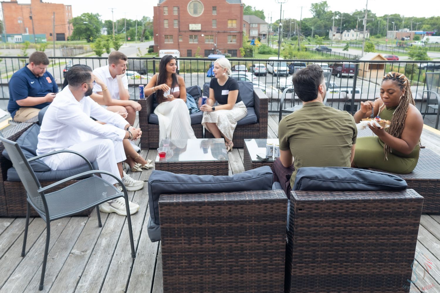 Group of people socializing on an outdoor rooftop patio with wicker furniture on a sunny day.