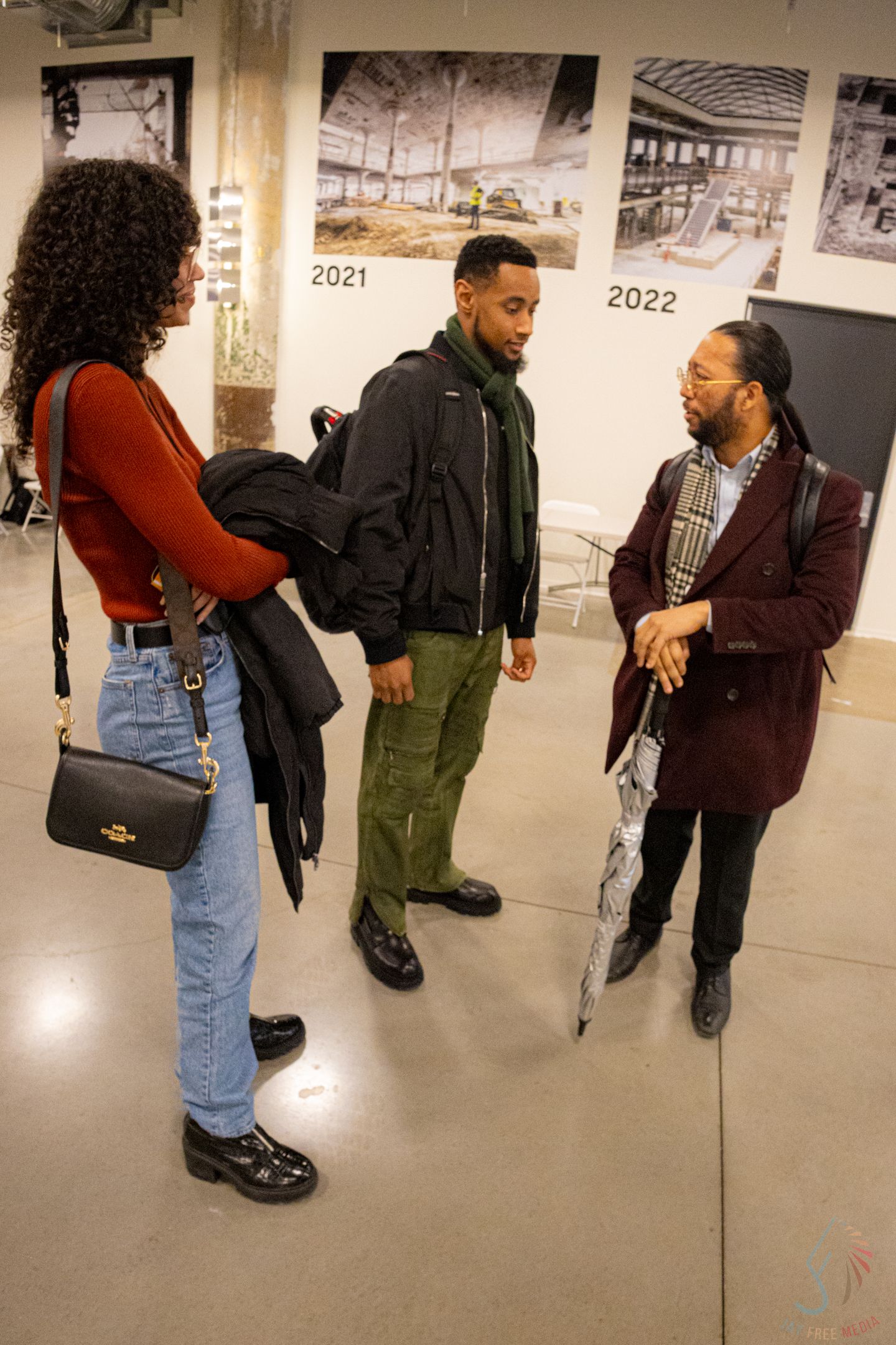 Three people engaged in conversation at an art gallery exhibition with timeline photographs displayed on the wall.
