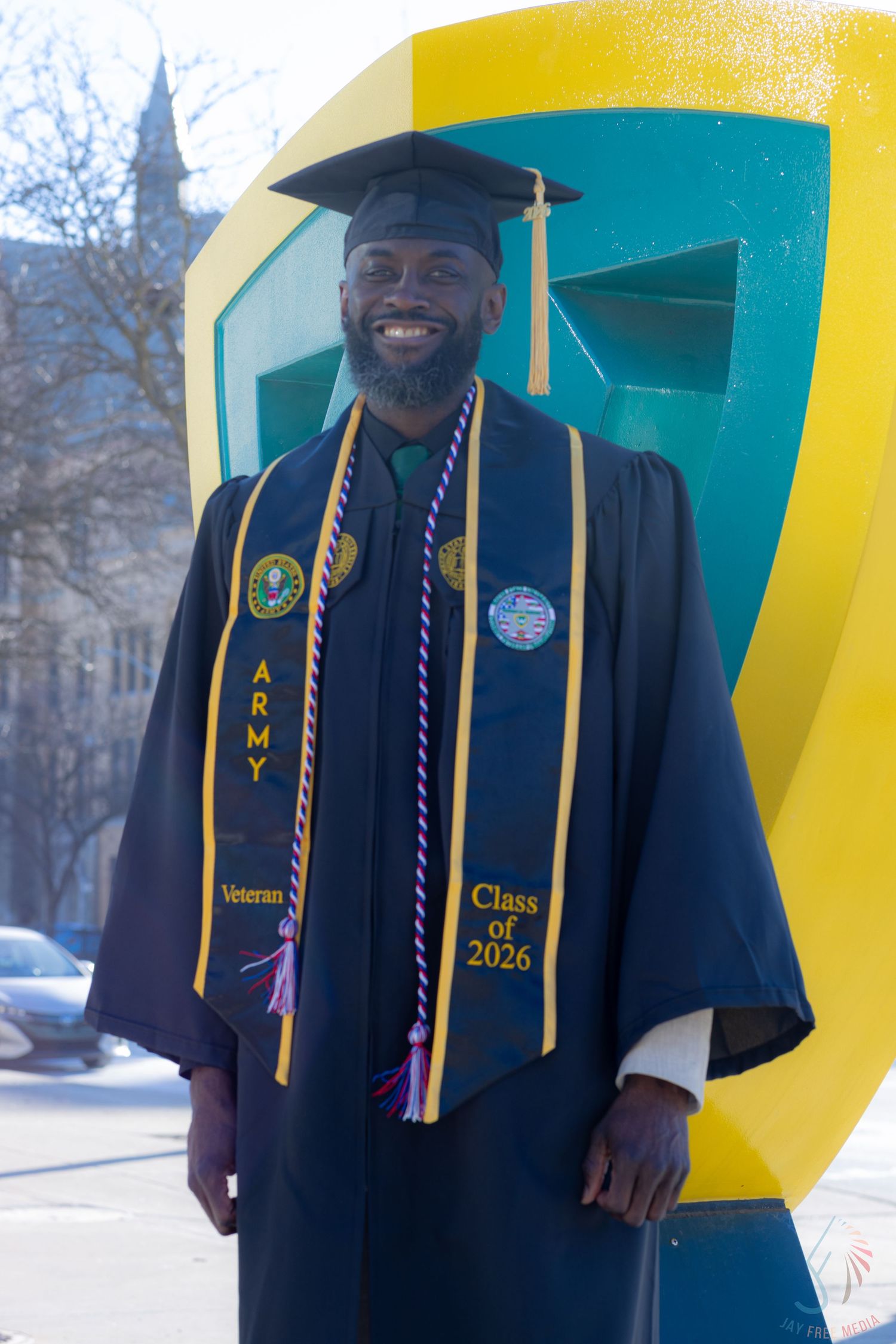 Army veteran smiles in graduation cap and gown with Class of 2026 stole, posing outdoors near colorful university sign.