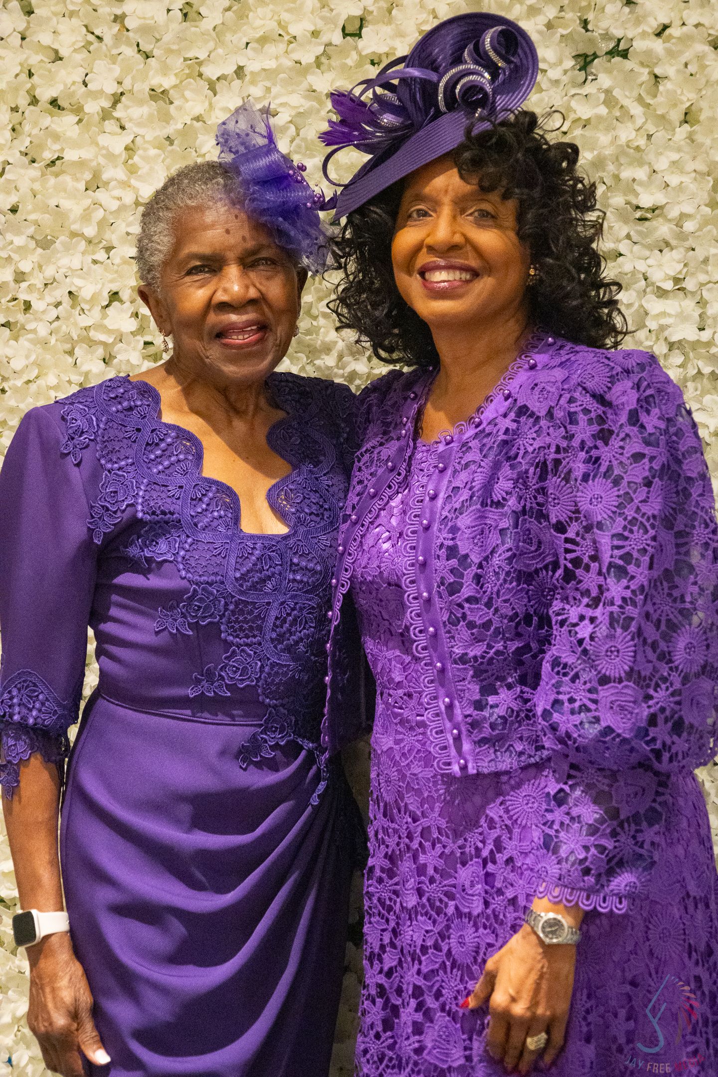 Two elegant women wearing matching purple lace dresses and decorative hats at a formal event.