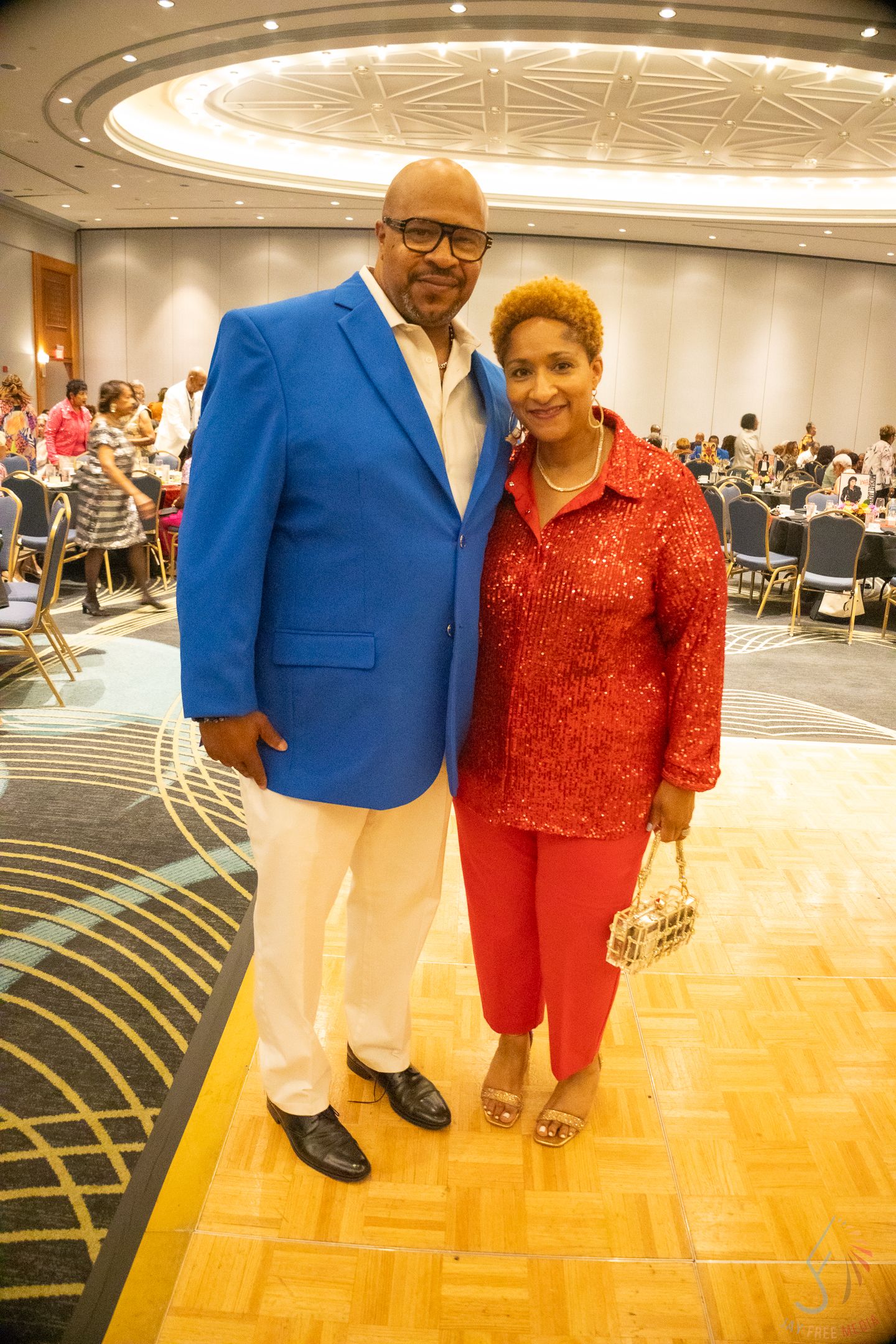 Couple posing at formal event, man in blue blazer and white pants, woman in red sequin top and pants.