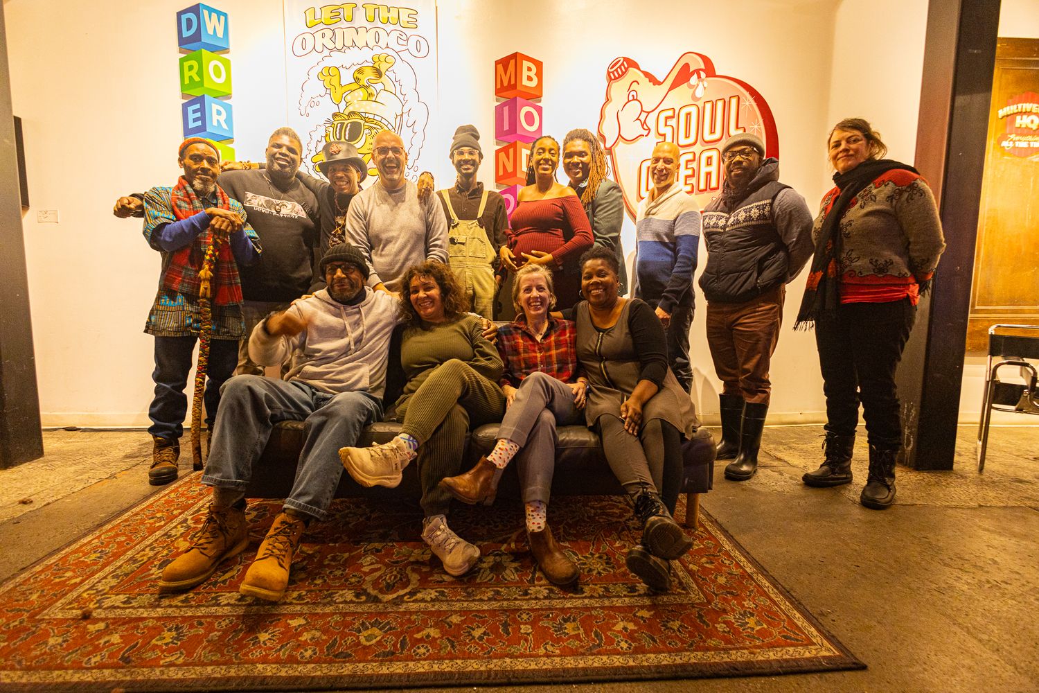 Diverse group of people posing together indoors near colorful signs, smiling for a group photo on a decorative rug.