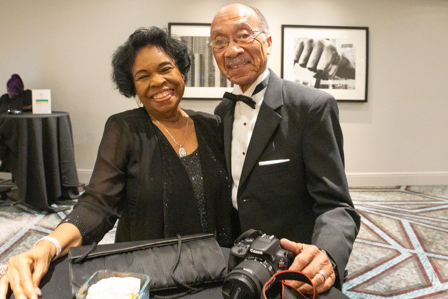 Elegant couple in formal attire smiling at a gala event, man holding a camera in a tuxedo.
