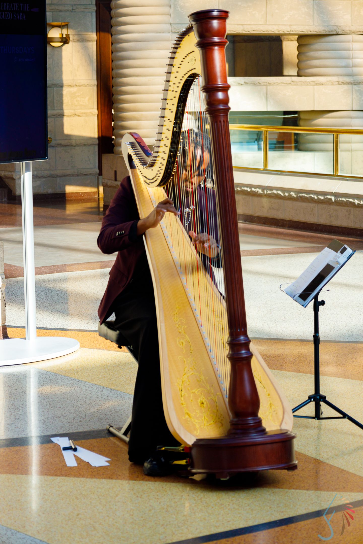 A musician plays a large wooden concert harp indoors, with sheet music on a stand nearby.
