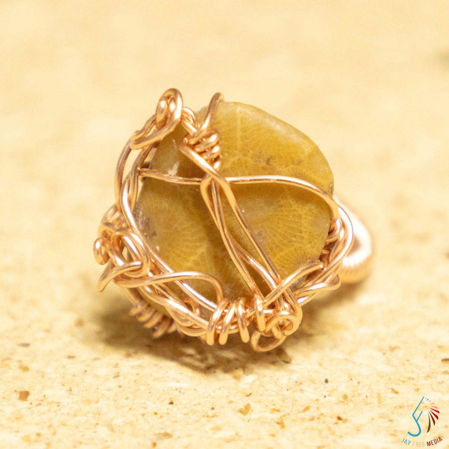 Handmade wire-wrapped ring featuring a golden yellow stone set in rose gold copper wire on a sandy background.