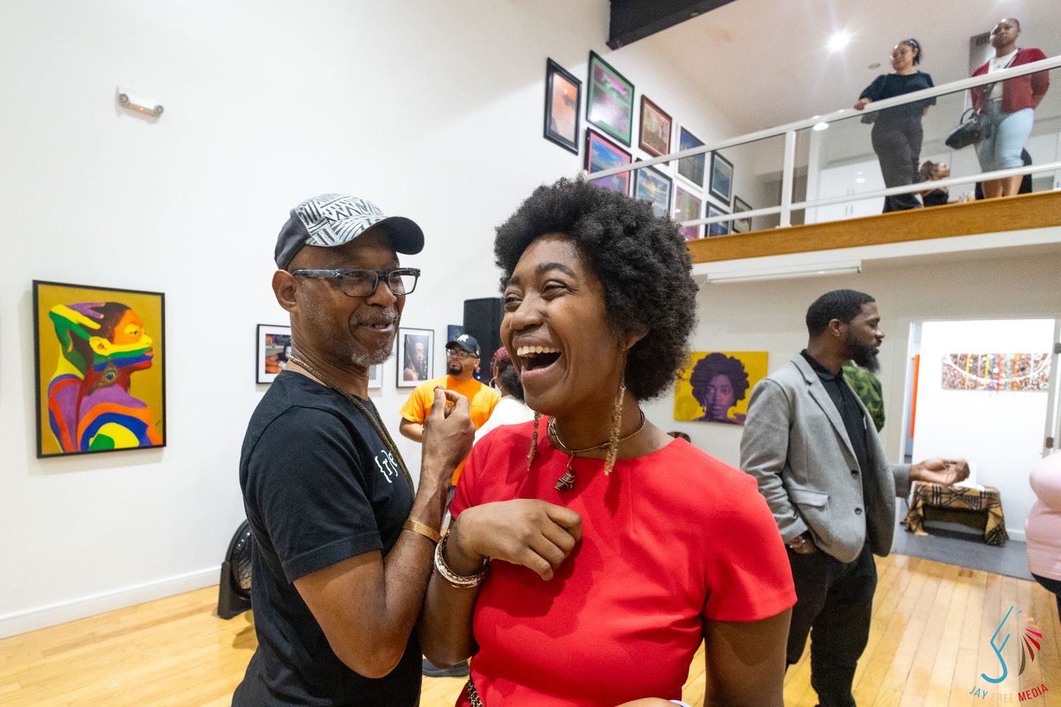 Two people laughing and socializing at an art gallery opening with colorful artwork displayed on white walls.