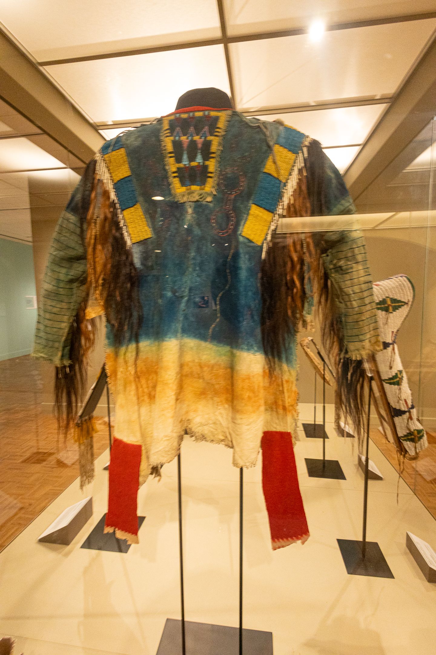 Colorful Native American fringed jacket with beadwork displayed in a glass museum case.