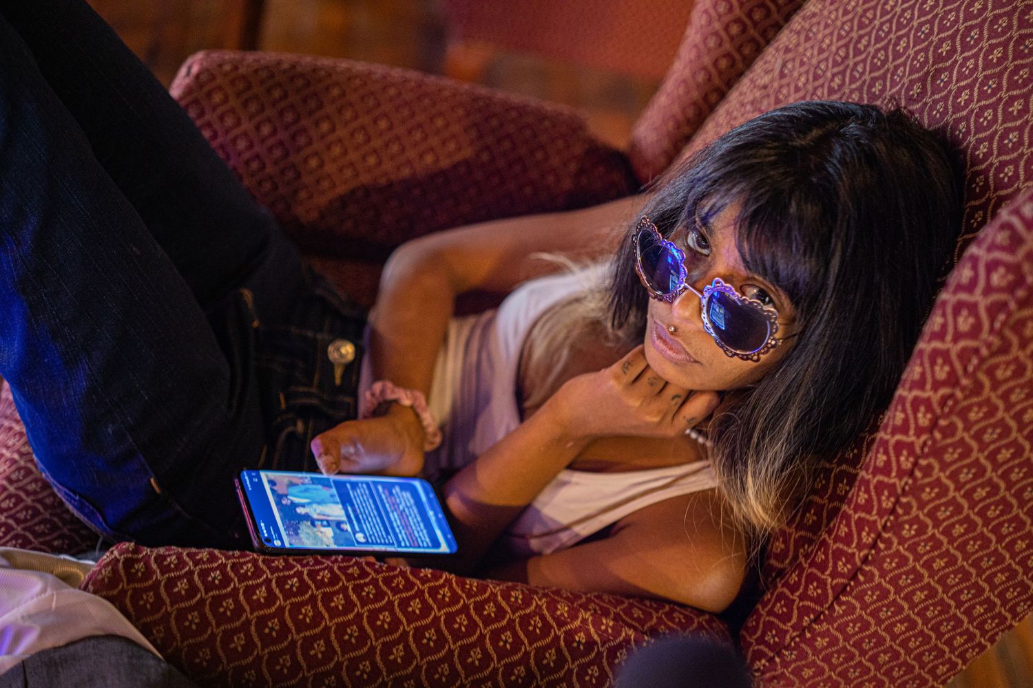 Young girl with sunglasses lying on a red armchair while using a tablet in dim lighting.