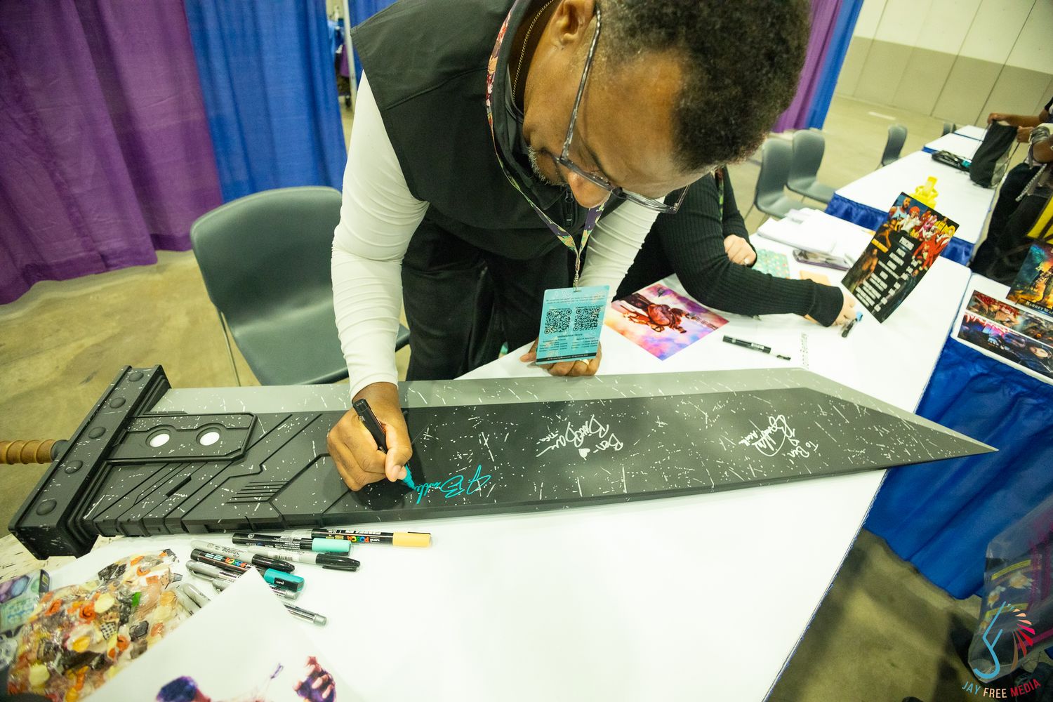 Person signing a large black sword prop at a convention table with colorful items displayed nearby.