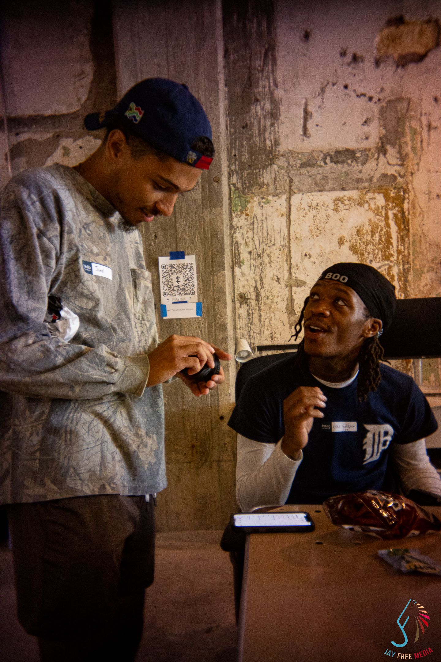 Two young men laugh together at a table in a dimly lit urban venue during an event.