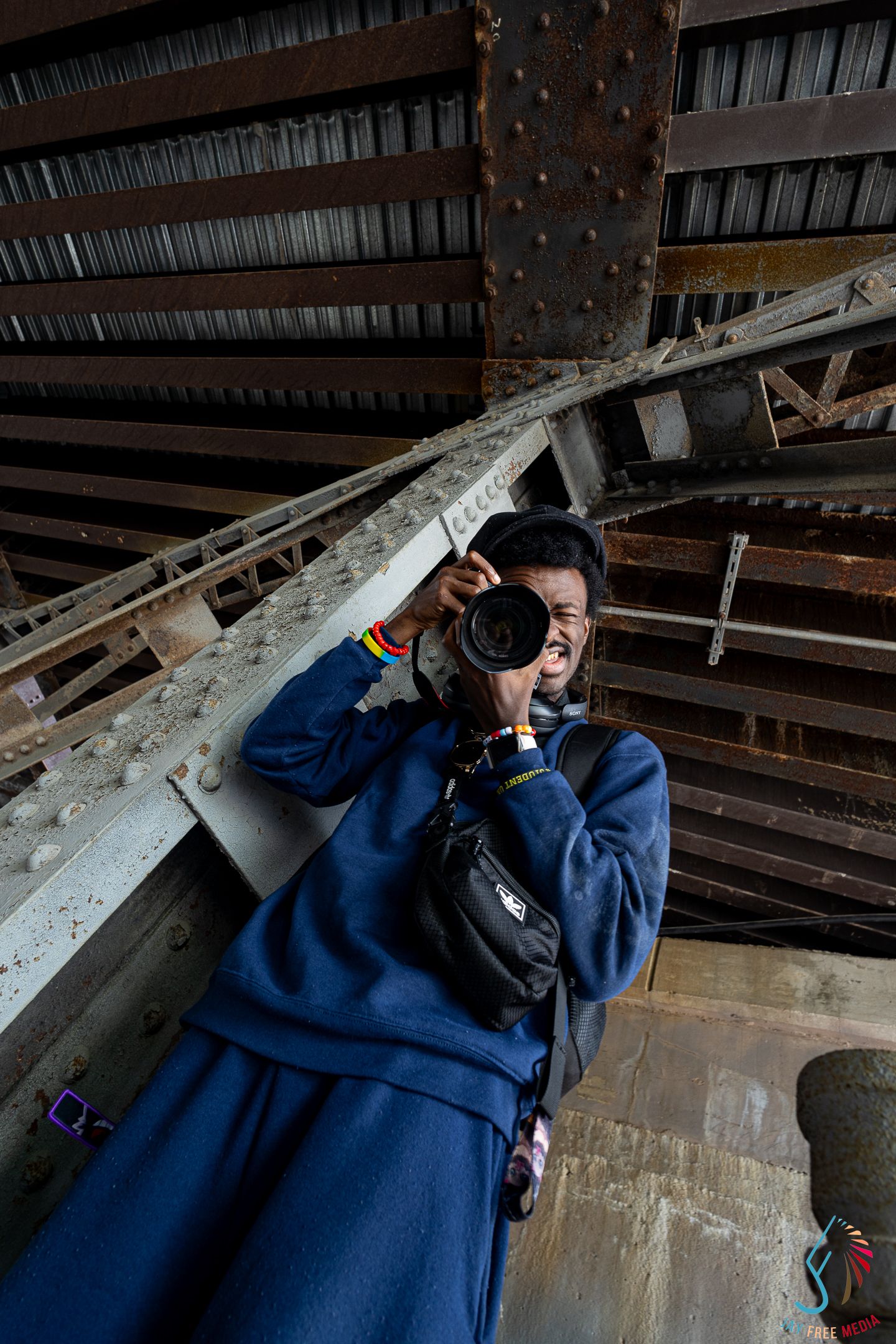 Photographer in blue hoodie captures shots under rusty industrial bridge structure.