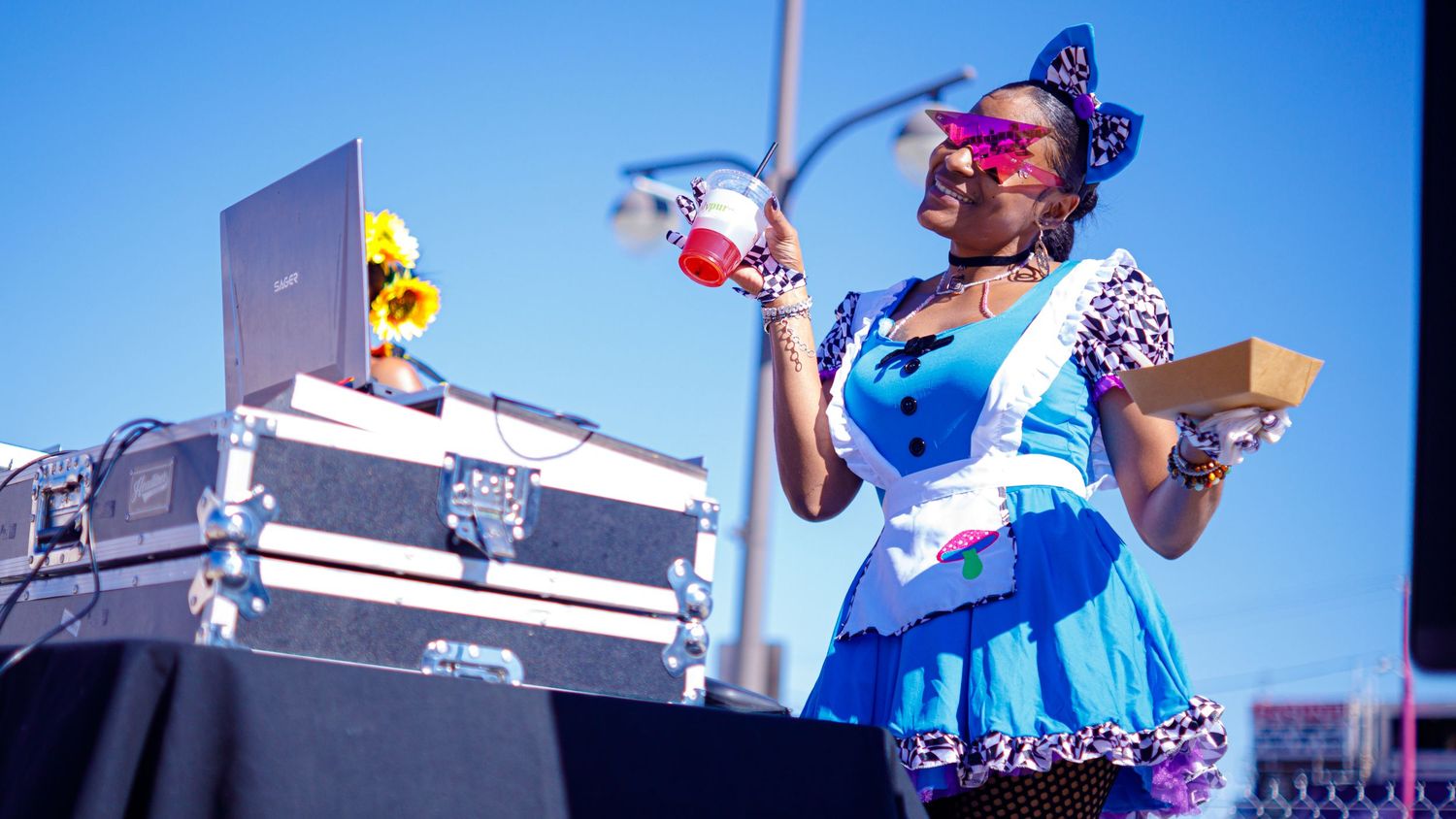 DJ in a blue dress performing on an outdoor stage under a sunny sky.