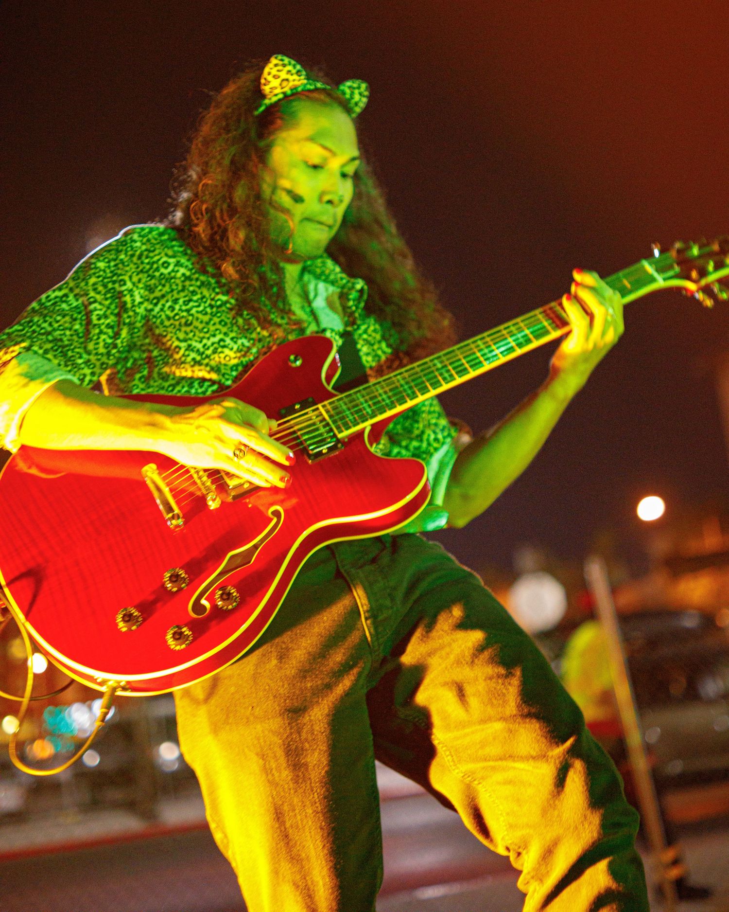 A guitarist with cat ears performs passionately on a red electric guitar under green stage lighting.