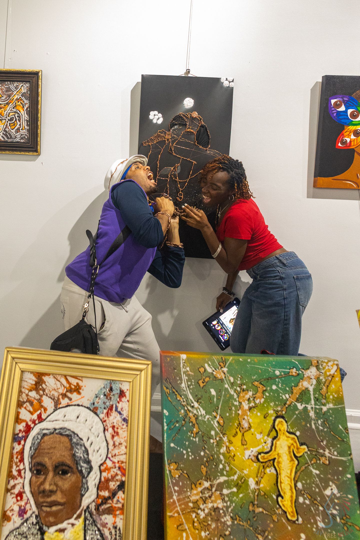 Two women examine a wire sculpture at an art gallery surrounded by colorful paintings on white walls.