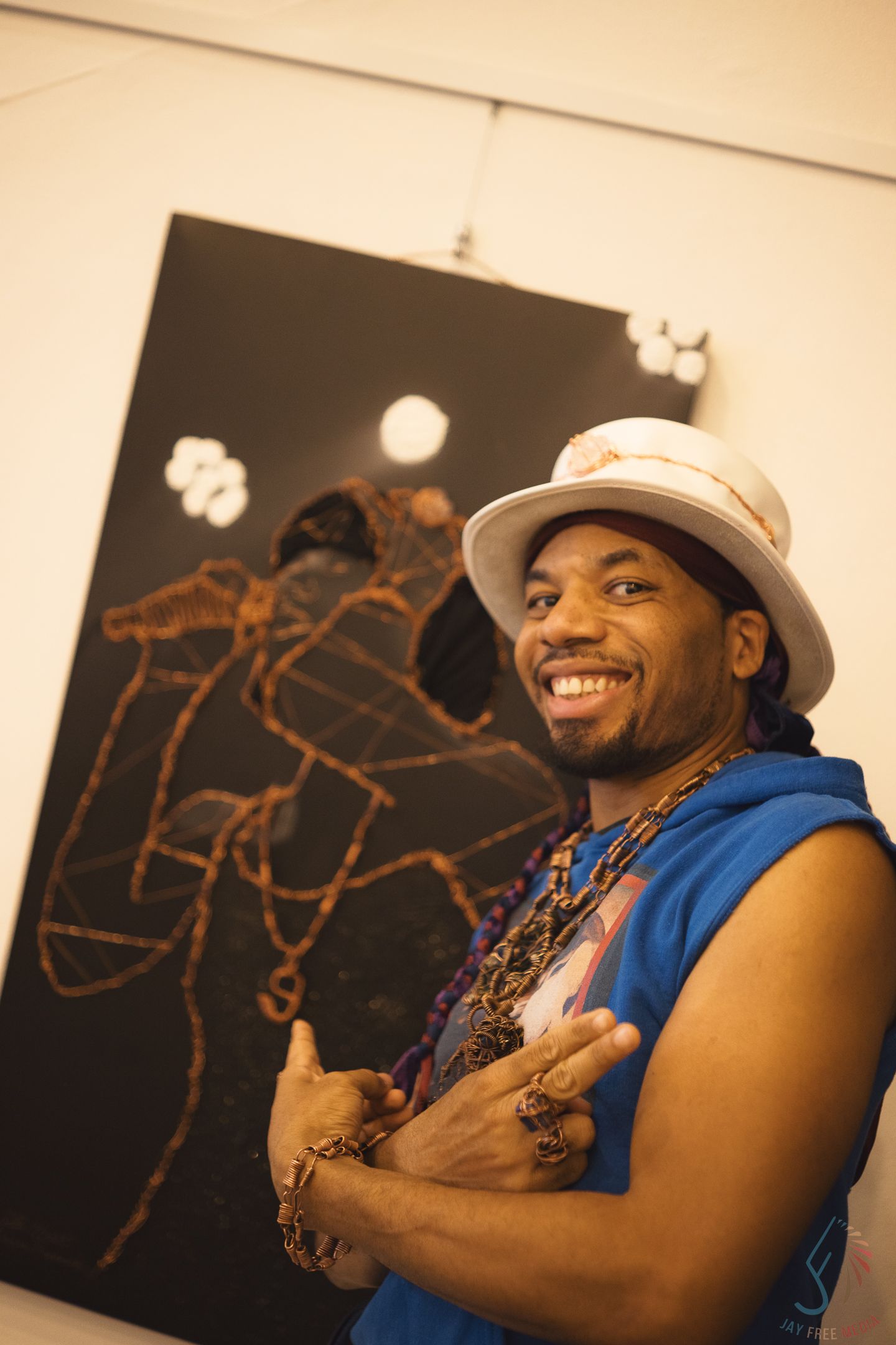 A smiling man in a white hat and blue vest poses with crossed arms before a dark abstract painting.