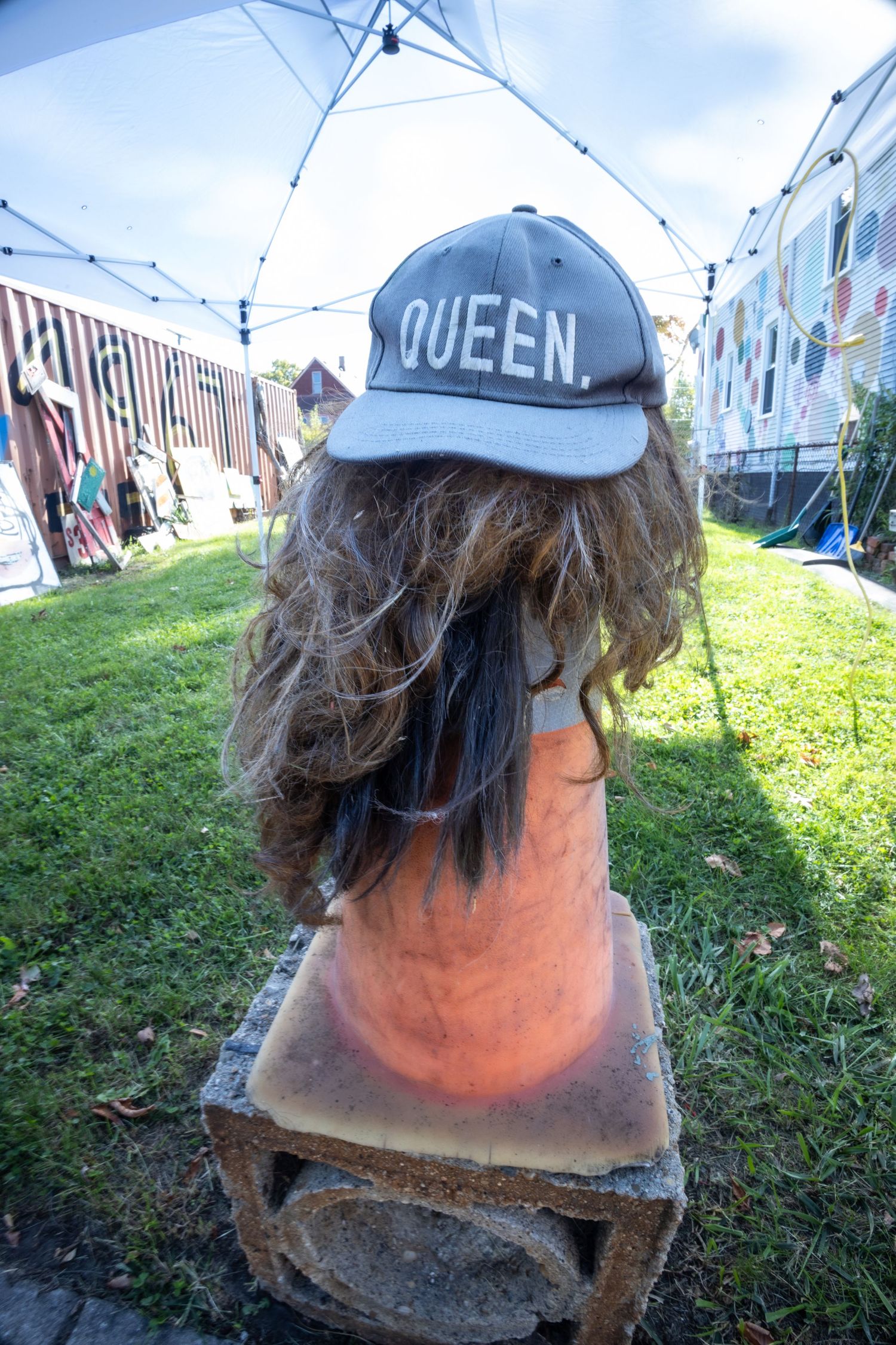 A wig with long brown hair displayed on a traffic cone wearing a gray cap that reads QUEEN at an outdoor market.