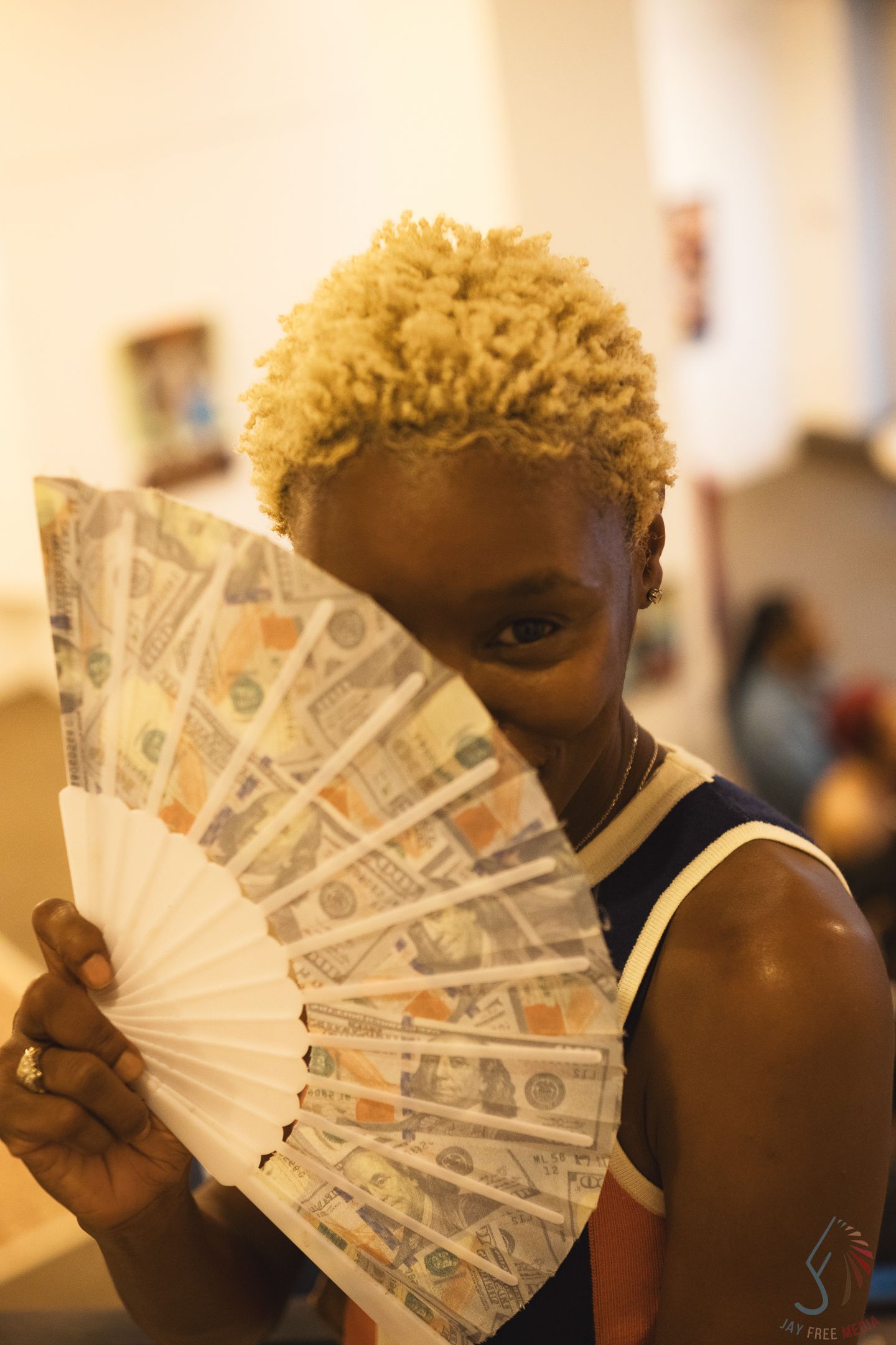 Black woman with blonde curly hair holding a decorative fan printed with dollar bills covering her face.