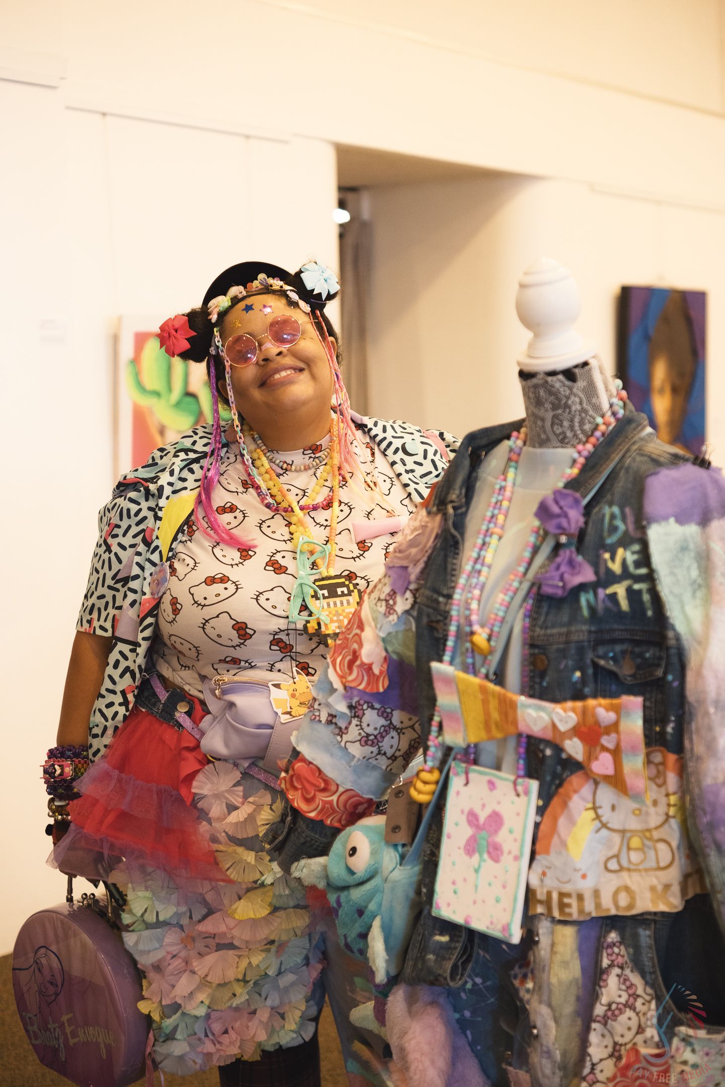 A colorfully dressed woman poses beside a Hello Kitty themed mannequin display at an art exhibition.
