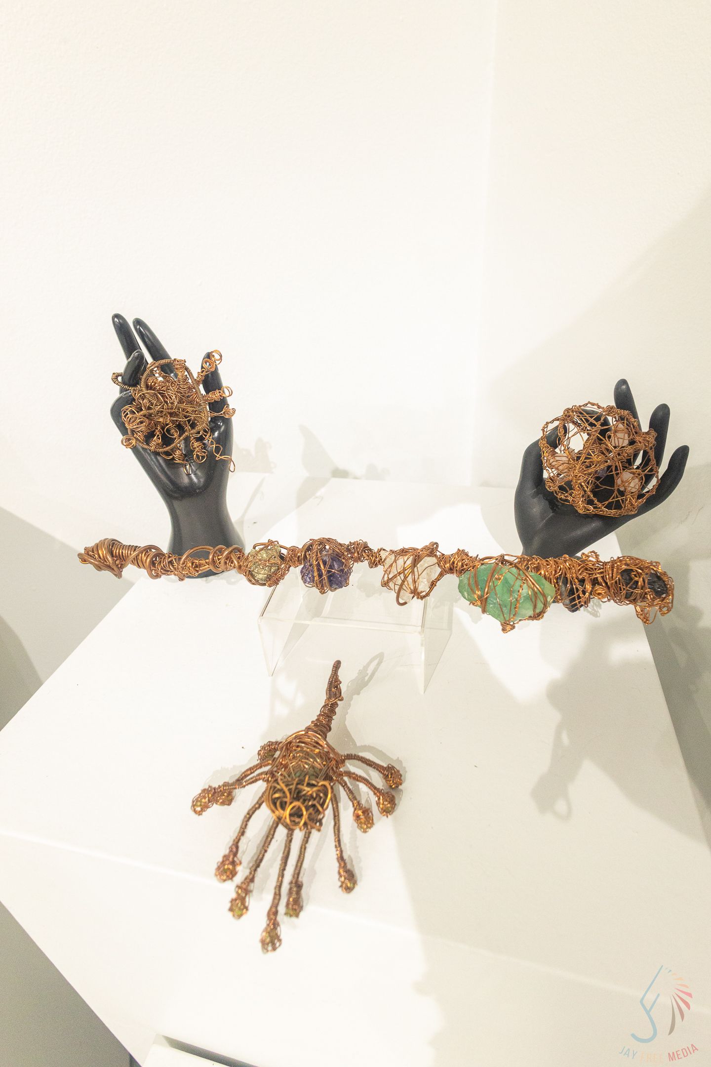 Black mannequin hands displaying copper wire-wrapped crystal jewelry pieces on a white geometric display stand.